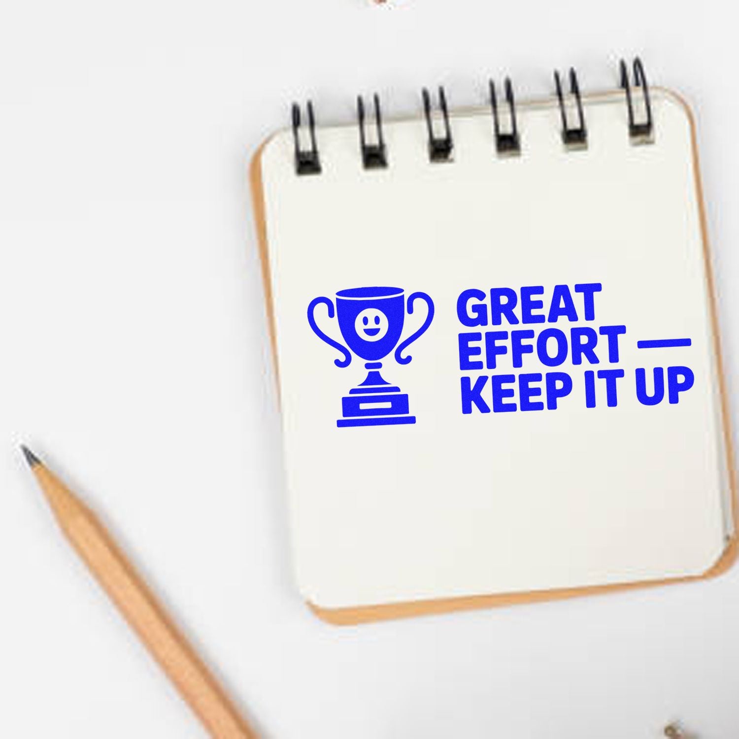 Blue motivational stamp seal with trophy icon and Great Effort Keep It Up on a white spiral notebook beside a pencil.