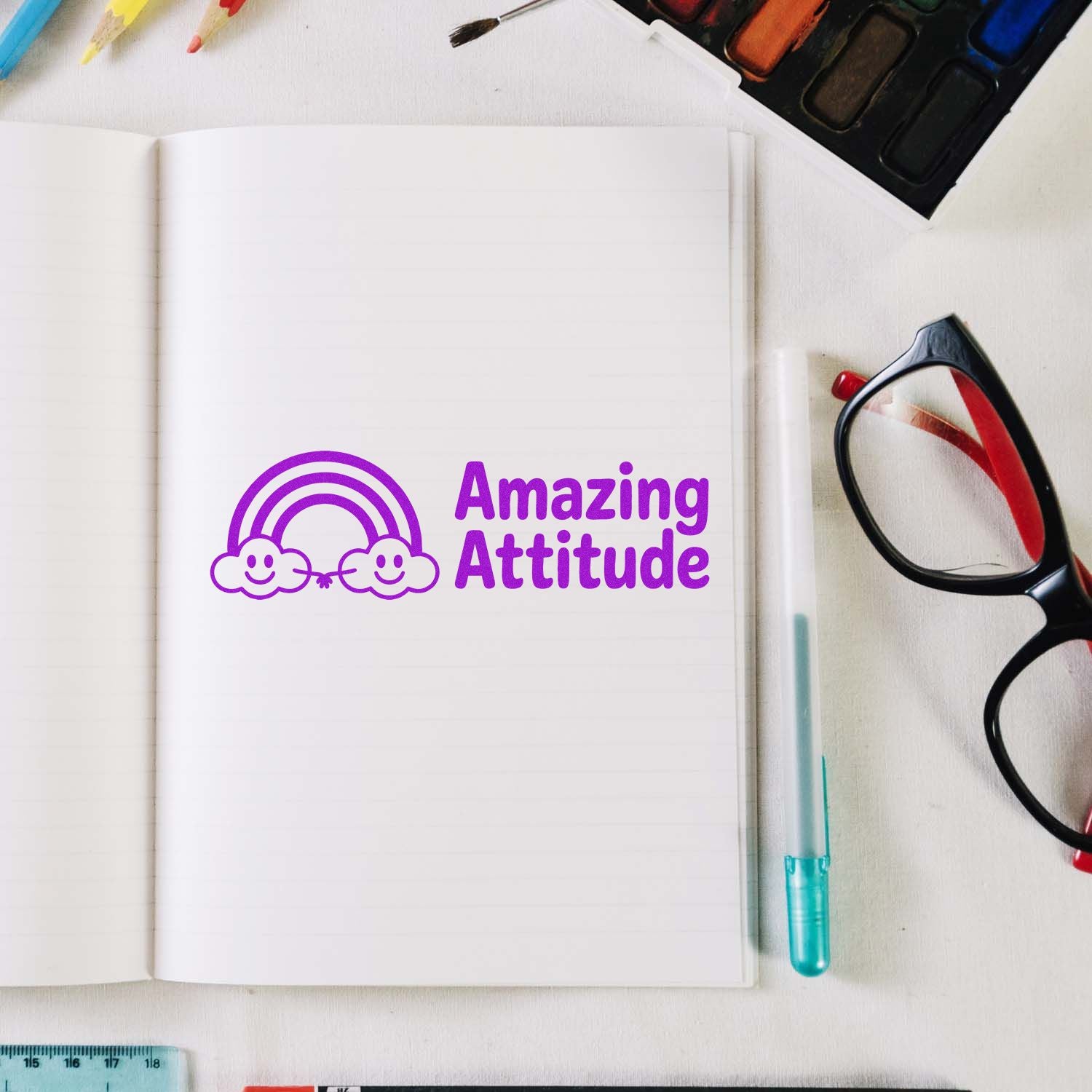 Purple self-inking stamp seal with smiling rainbow clouds and Amazing Attitude on a lined notebook, beside glasses.