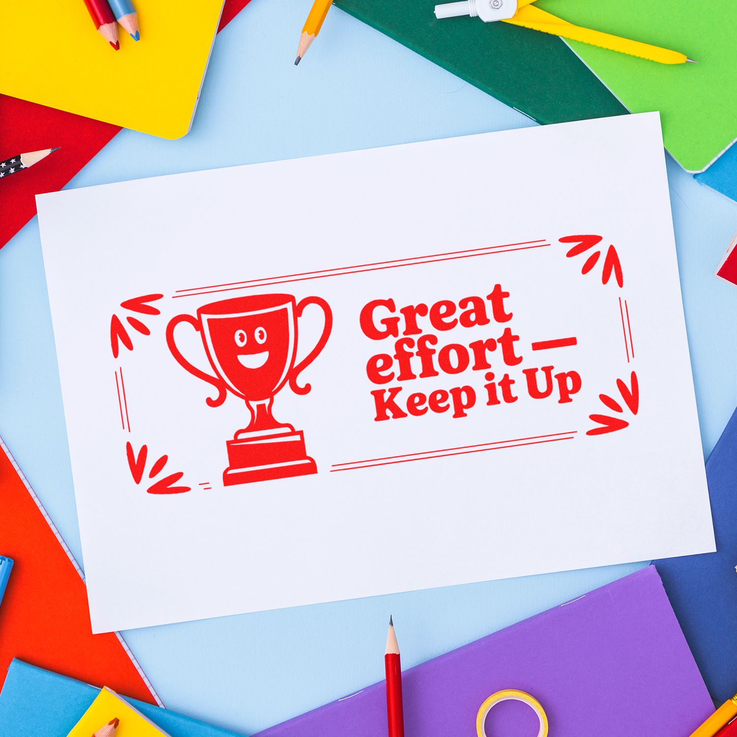 Red self-inking stamp with trophy and Great effort Keep it Up on white paper, framed by colorful pencils and notebooks.