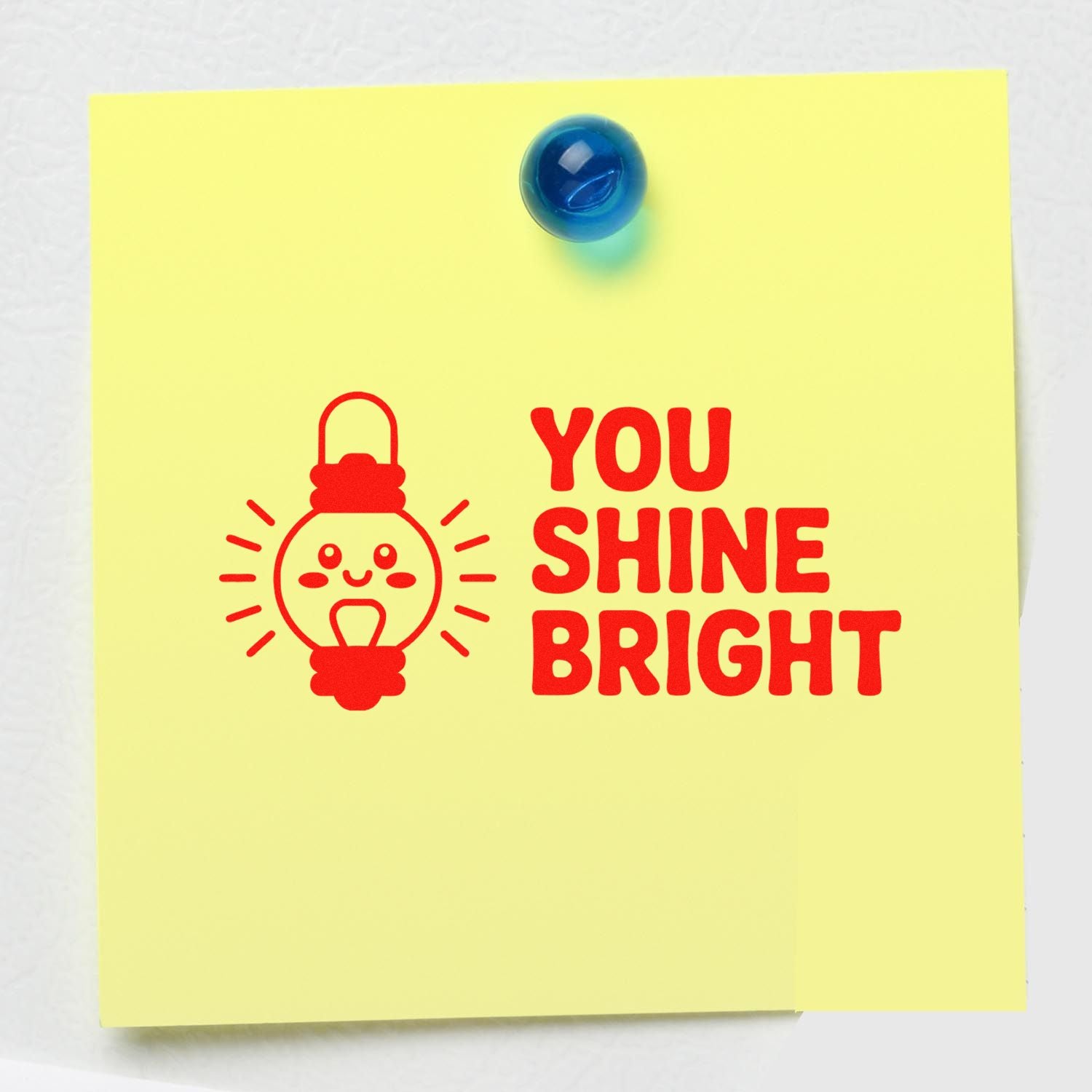 Red rubber stamp seal reading You Shine Bright with smiling light bulb on yellow note, pinned by a blue pushpin.