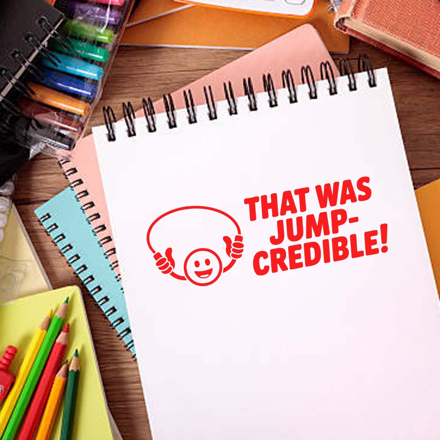 Red stamp seal reading THAT WAS JUMP-CREDIBLE! on a white spiral notebook, with a smiling thumbs-up icon and colorful stationery.