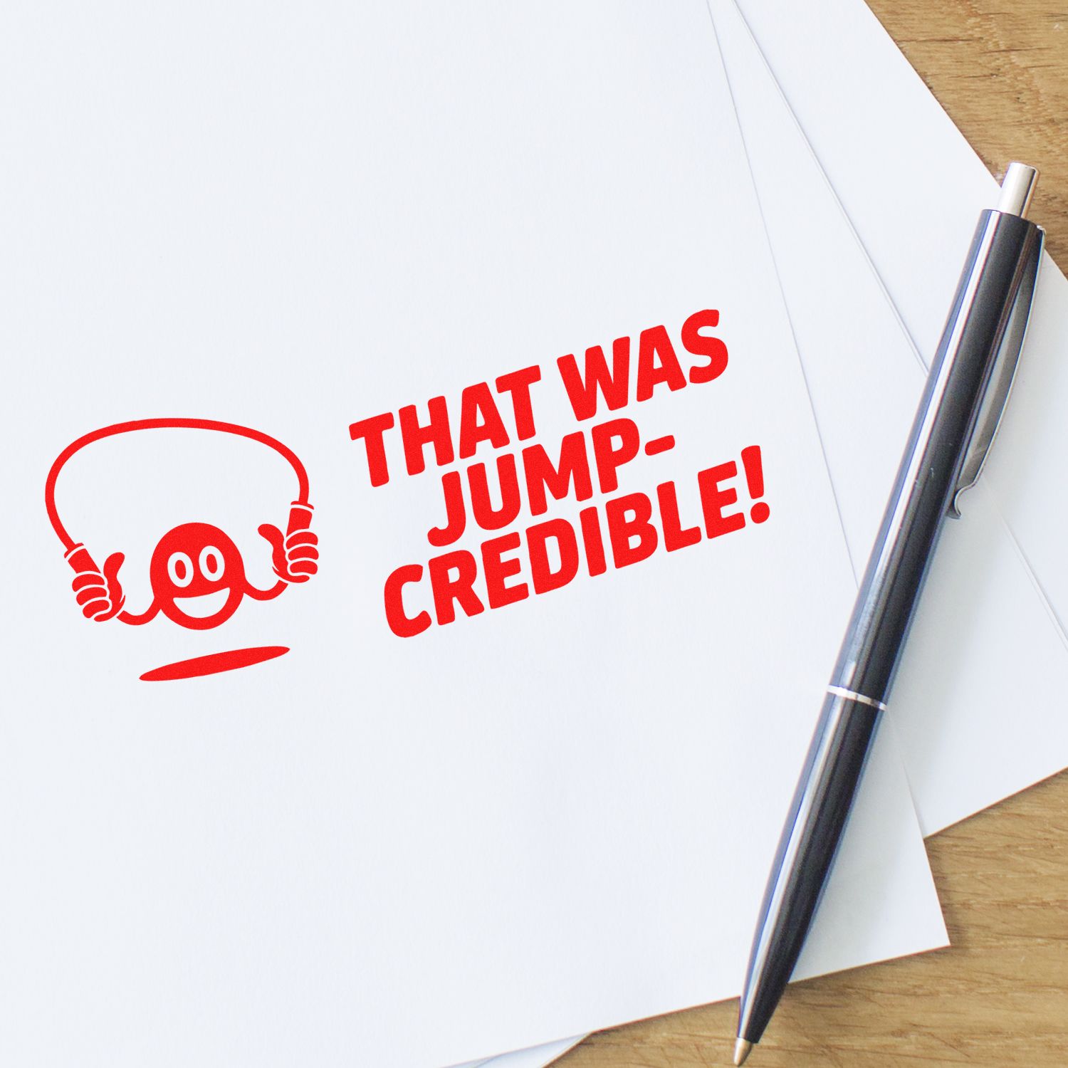 Red stamp and seal reading THAT WAS JUMP-CREDIBLE! with mascot logo on white paper, beside a black pen on a wood desk.
