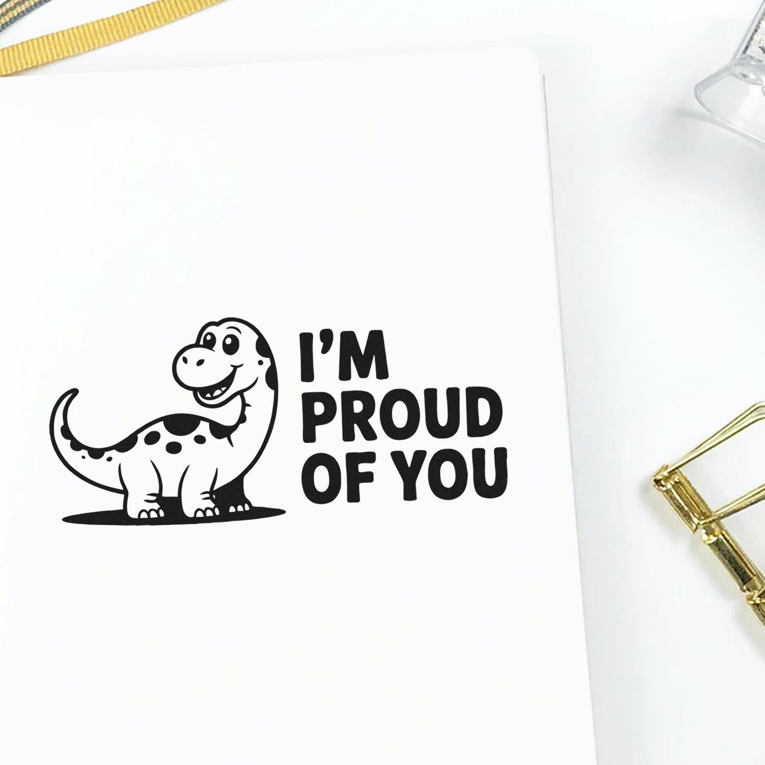 Black dinosaur stamp reading IM PROUD OF YOU on white paper, with a gold seal clip.
