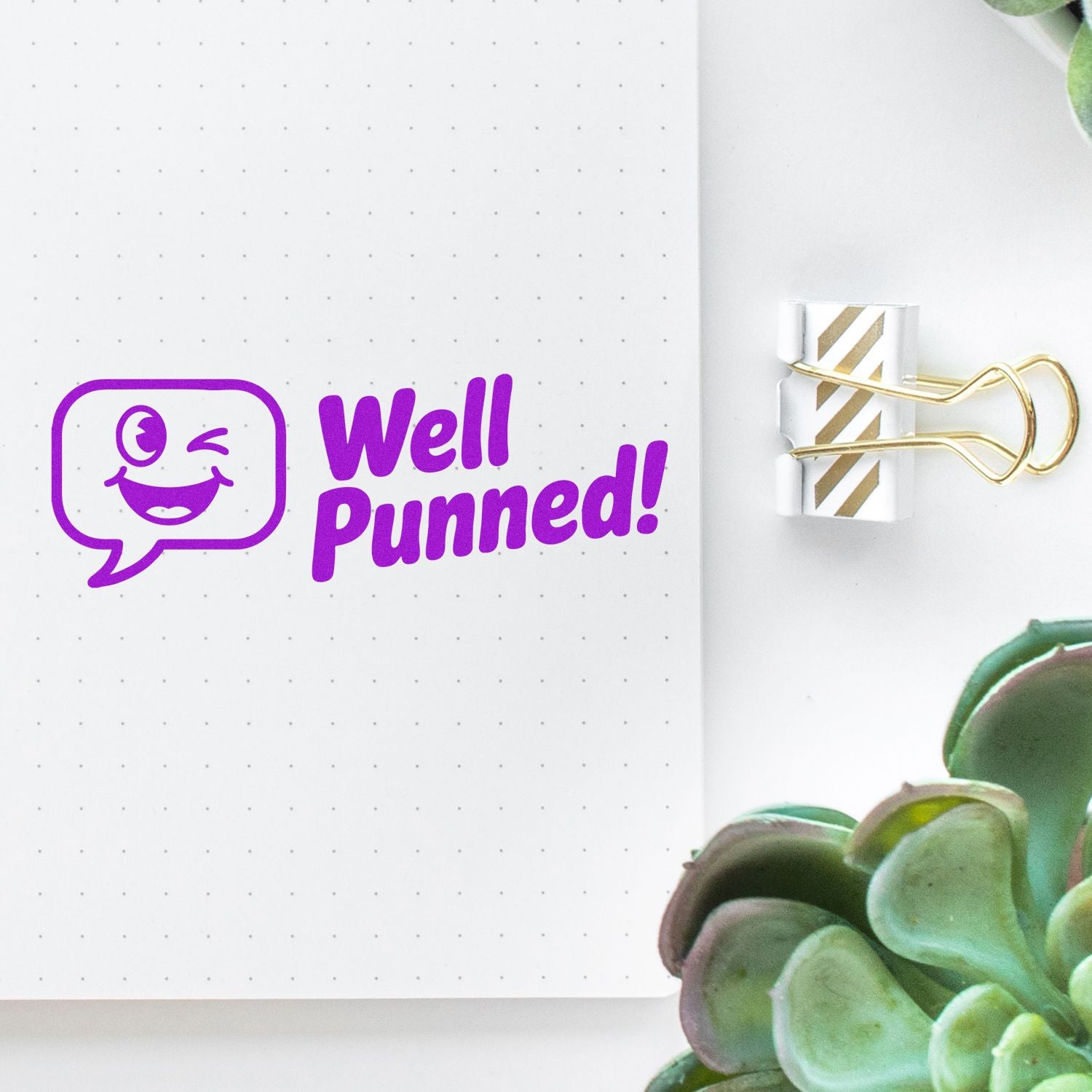 Purple Well Punned! stamp and gold-striped seal clip on dotted notebook page with winking face icon.