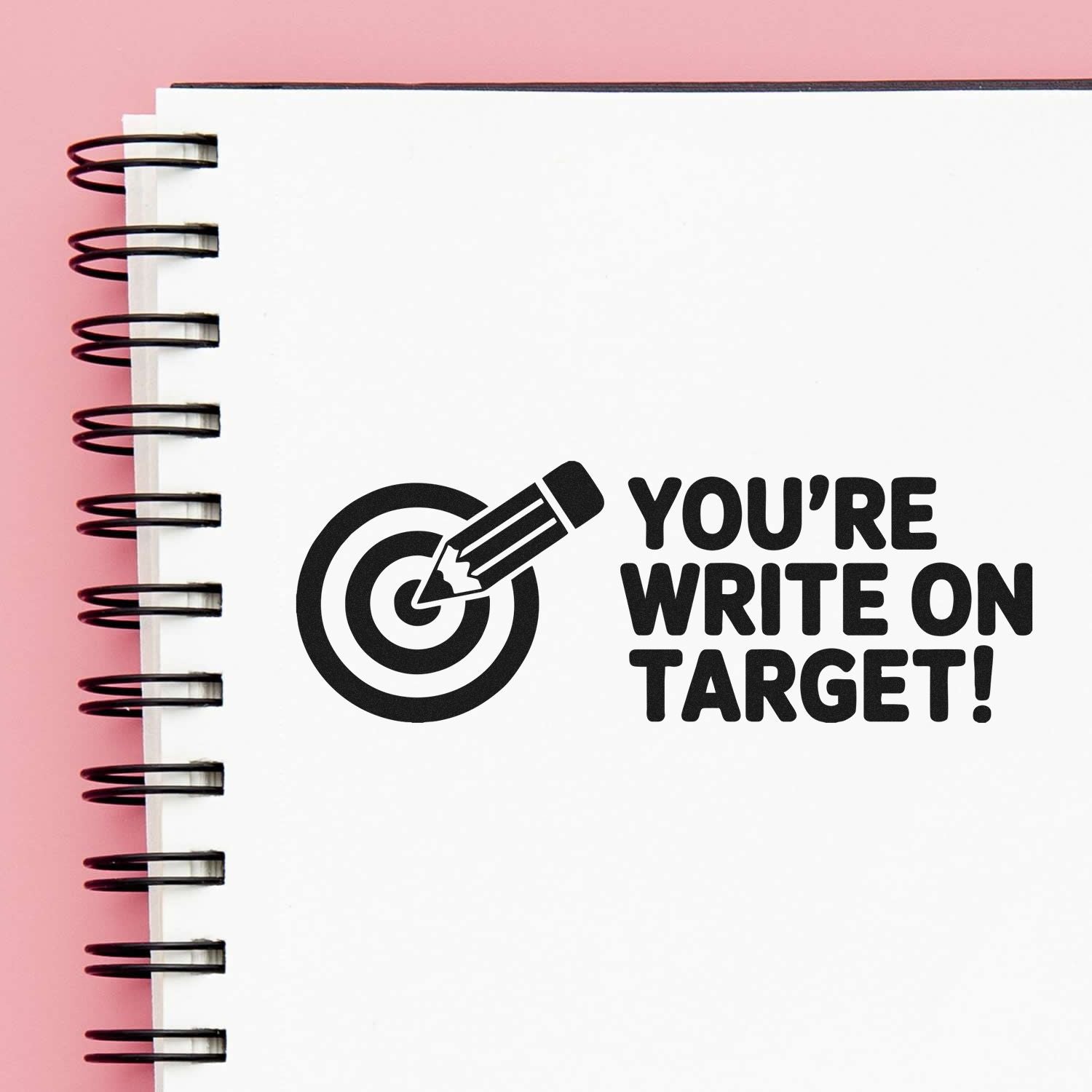 Black stamp-style target seal with pencil and YOURE WRITE ON TARGET! on a white spiral notebook against a pink background.