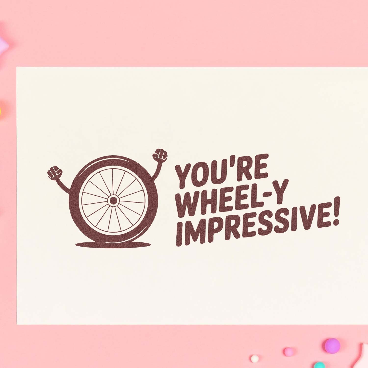 Burgundy rubber stamp seal of a cheering wheel with YOURE WHEEL-Y IMPRESSIVE! on cream card against pink background