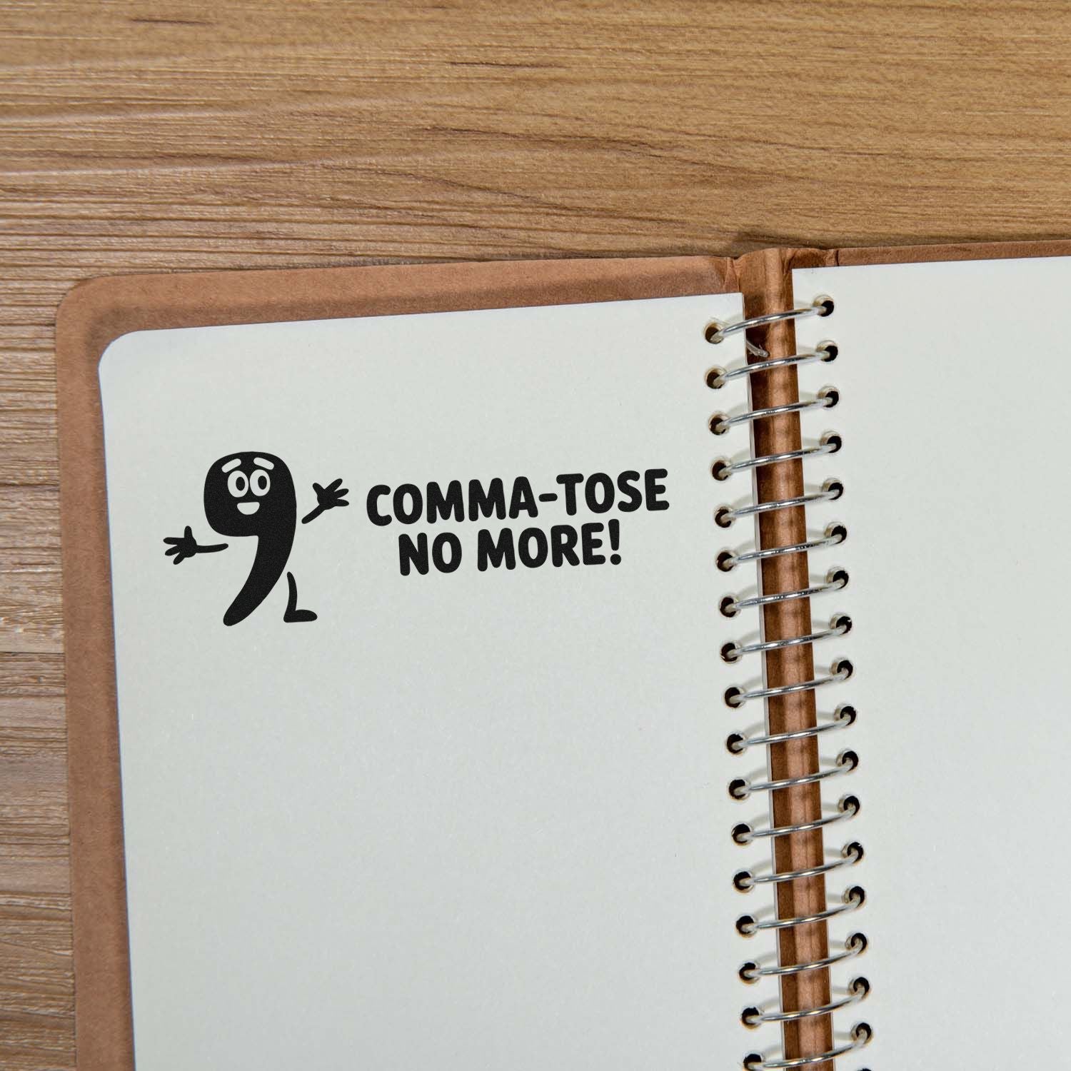 Black stamp of a comma character with COMMA-TOSE NO MORE! on a white spiral notebook, brown cover, wood desk.