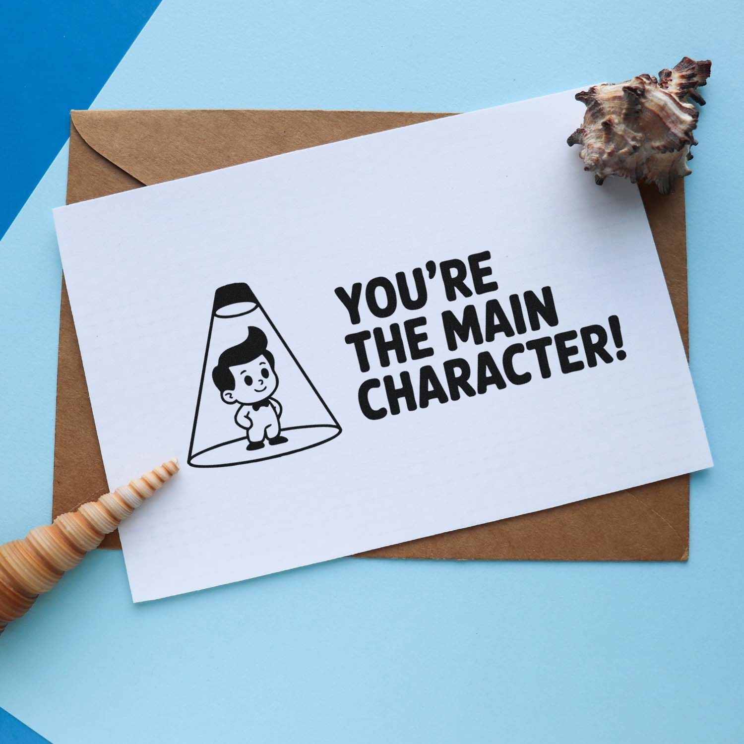 Black stamp and shell seal on white card reads Youre the main character! with brown envelope on blue background.
