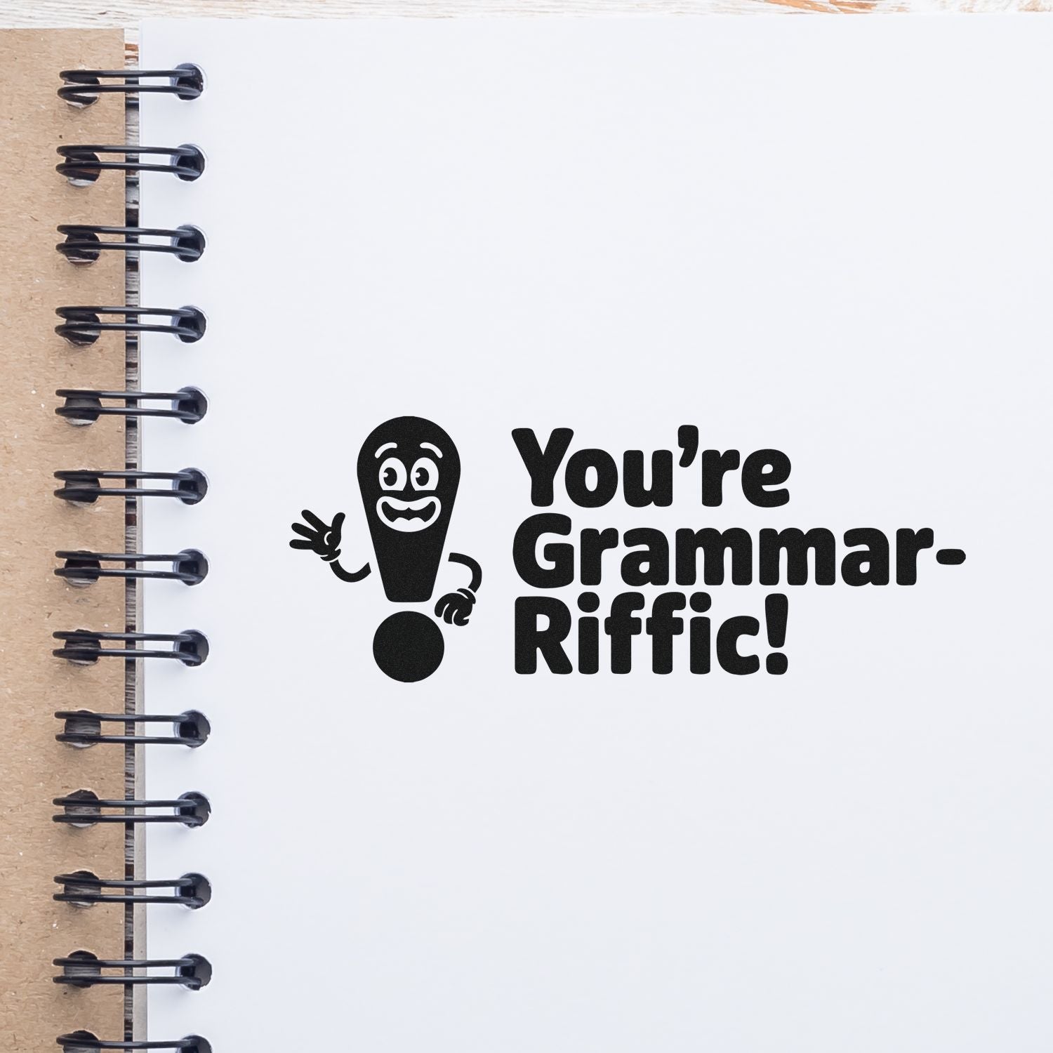 Black grammar-riffic stamp impression on a white notebook page, with a smiling exclamation mark and black spiral binding.