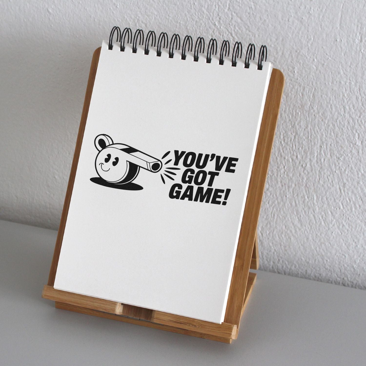 Black YOUVE GOT GAME! stamp on a white notepad in a wooden stand, featuring a playful whistle graphic.
