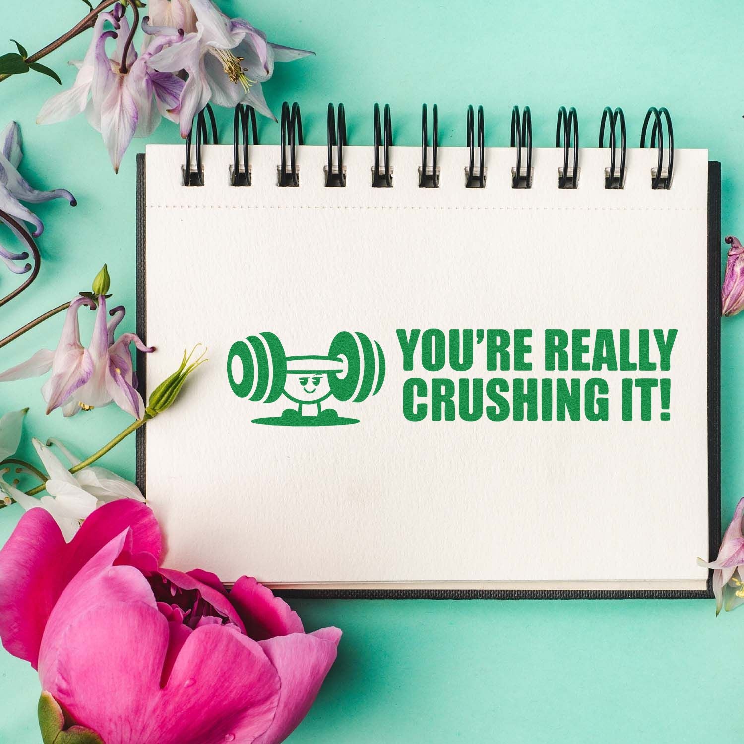 Teal background with spiral notebook, green stamp and seal reading Youre really crushing it! framed by pink and purple flowers