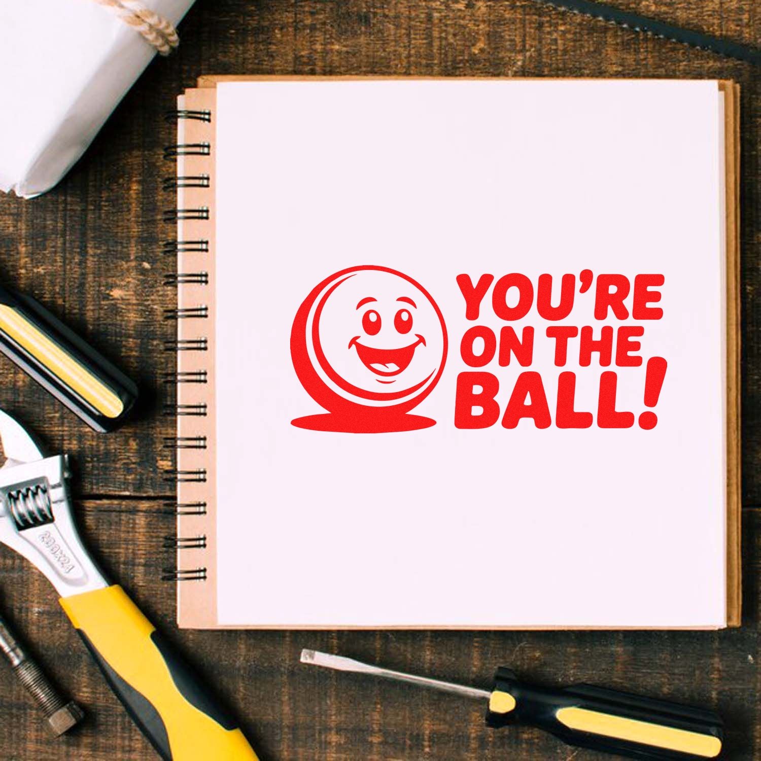 Red rubber stamp seal on a white notebook reads YOURE ON THE BALL! with a smiling ball icon on rustic wood, surrounded by tools.