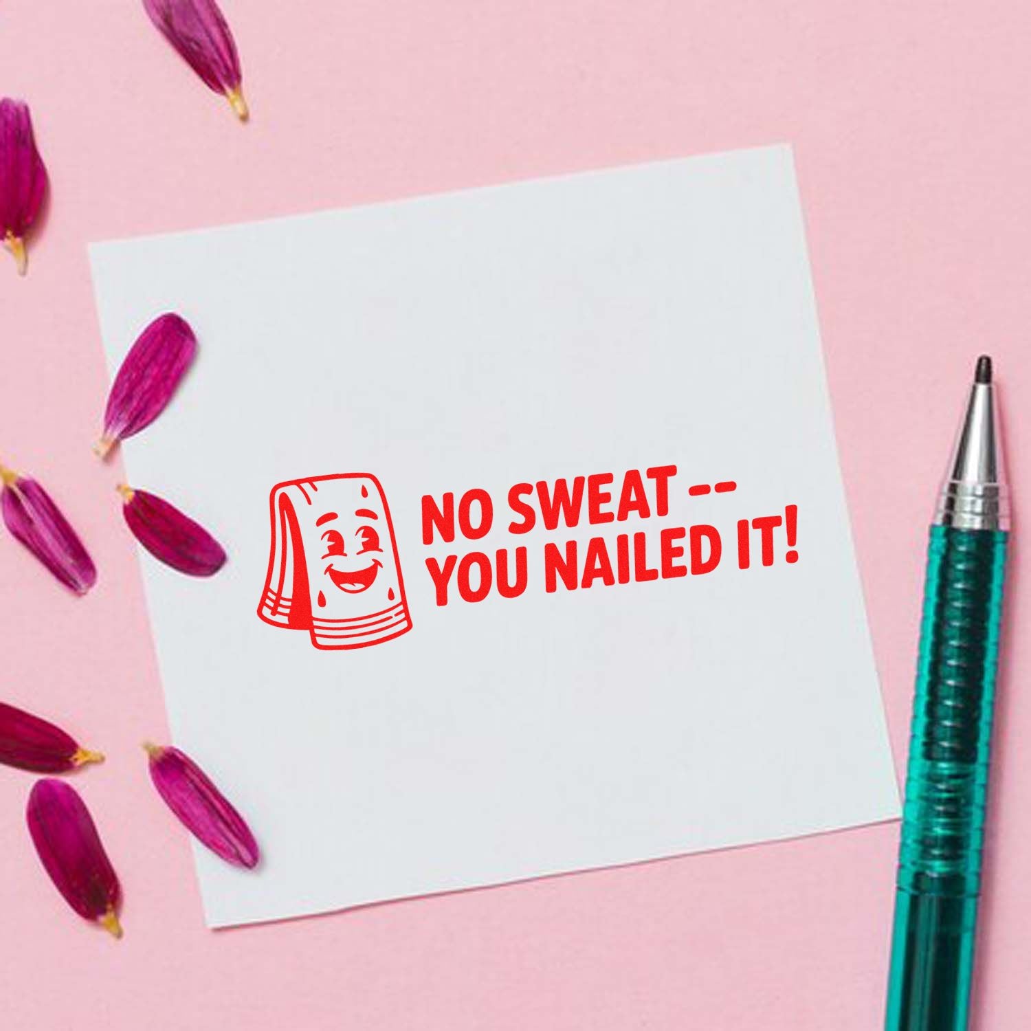 Red self-inking stamp seal reading No SweatYou Nailed It! on white card, with pink petals and a teal pen on pink background.