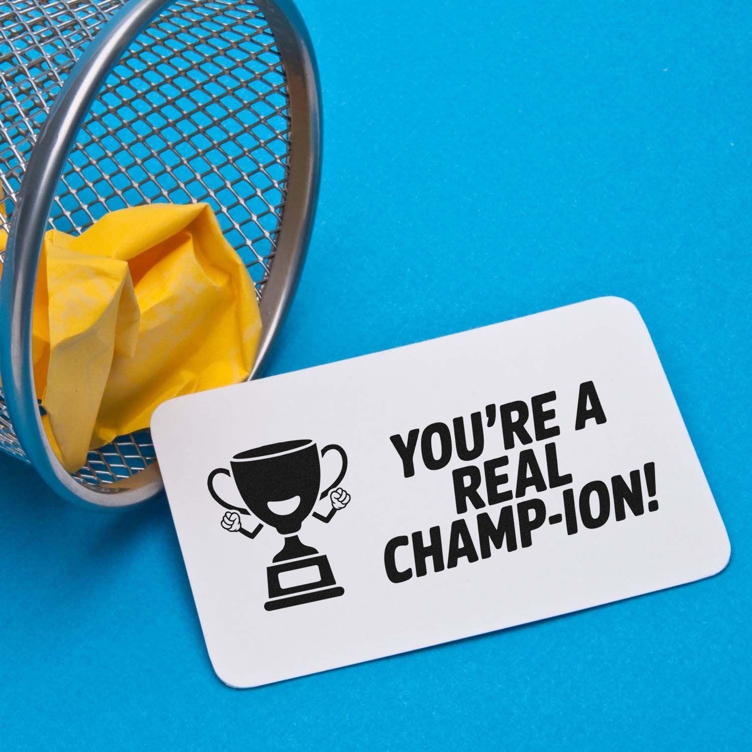 White stamp/seal card with black trophy and Youre a real champ-ion! on bright blue background beside yellow paper basket