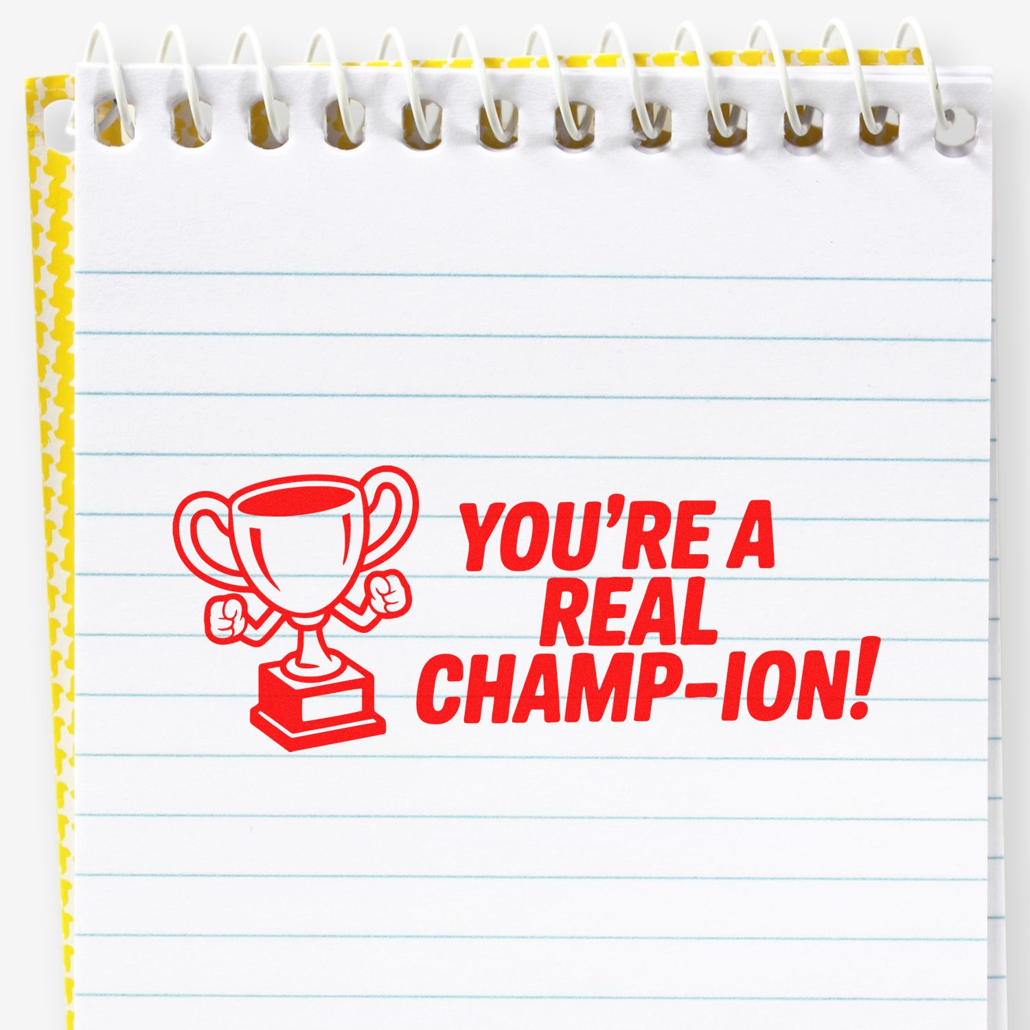 Red rubber stamp on lined notepad says YOURE A REAL CHAMP-ION! with trophy icon, yellow border, and spiral binding.