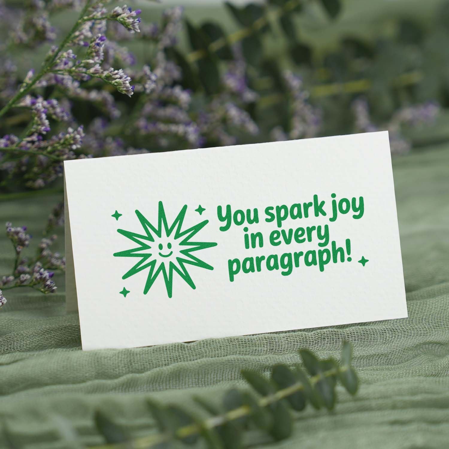 Green custom stamp and seal on white card with starburst and You spark joy in every paragraph! text, among purple flowers.