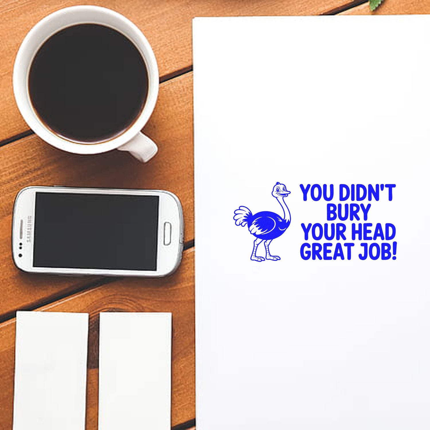 Blue ostrich stamp and seal with You didnt bury your head great job! on white paper, beside phone, coffee, and pads.