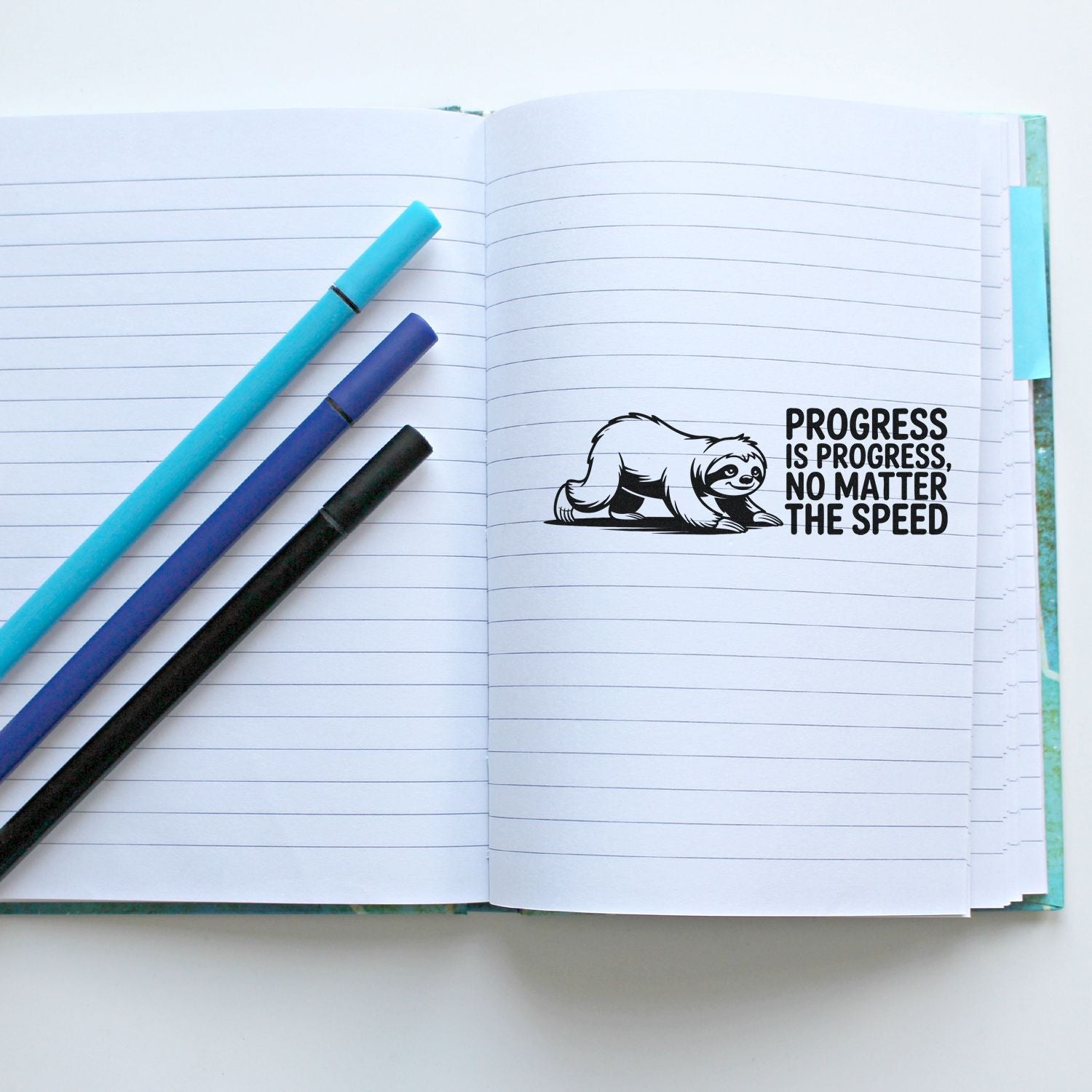 Open lined notebook with turquoise, blue and black pens beside a black sloth progress stamp seal.