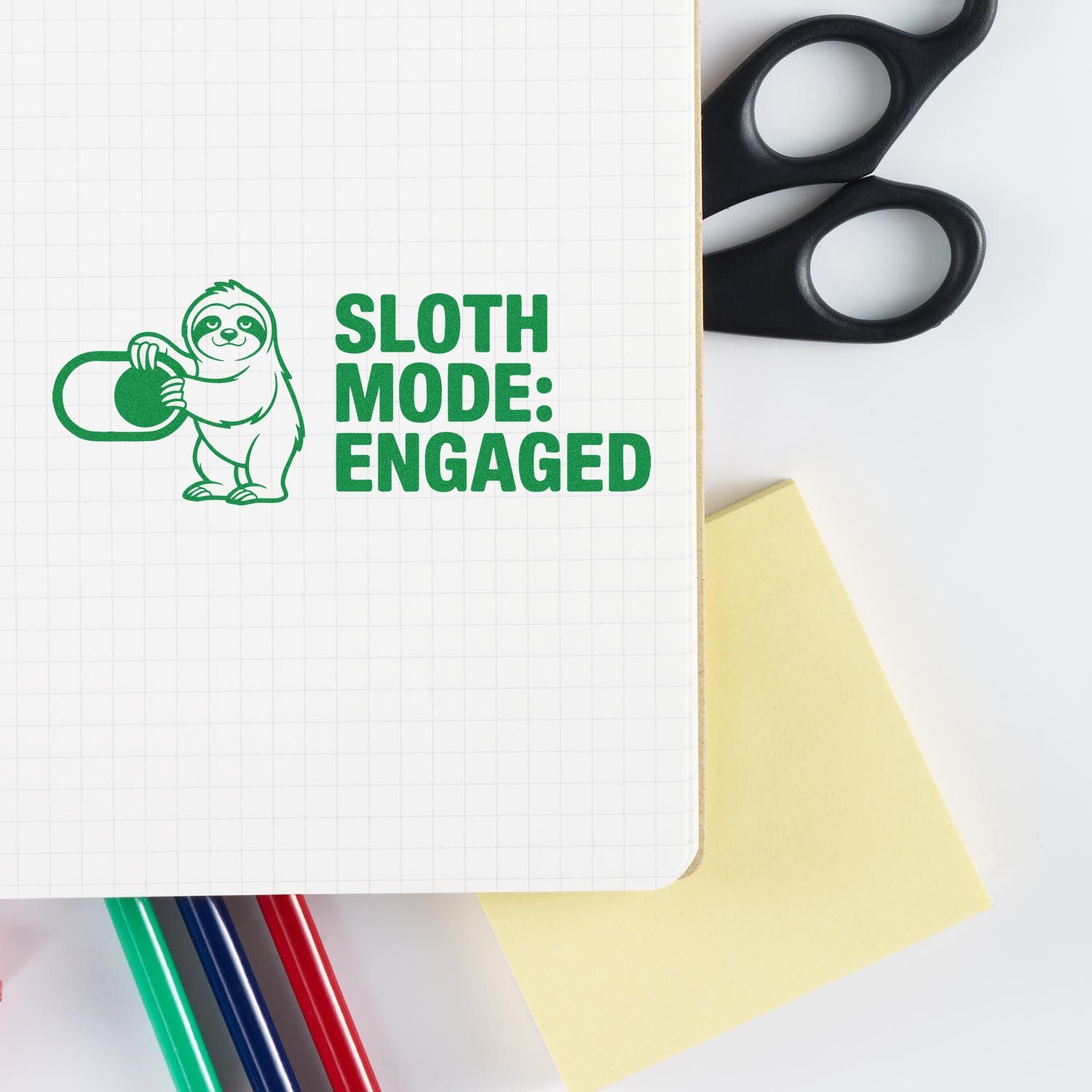 Green custom rubber stamp with sloth and Sloth Mode: Engaged on grid paper, plus black scissors, sticky note and colored pens.