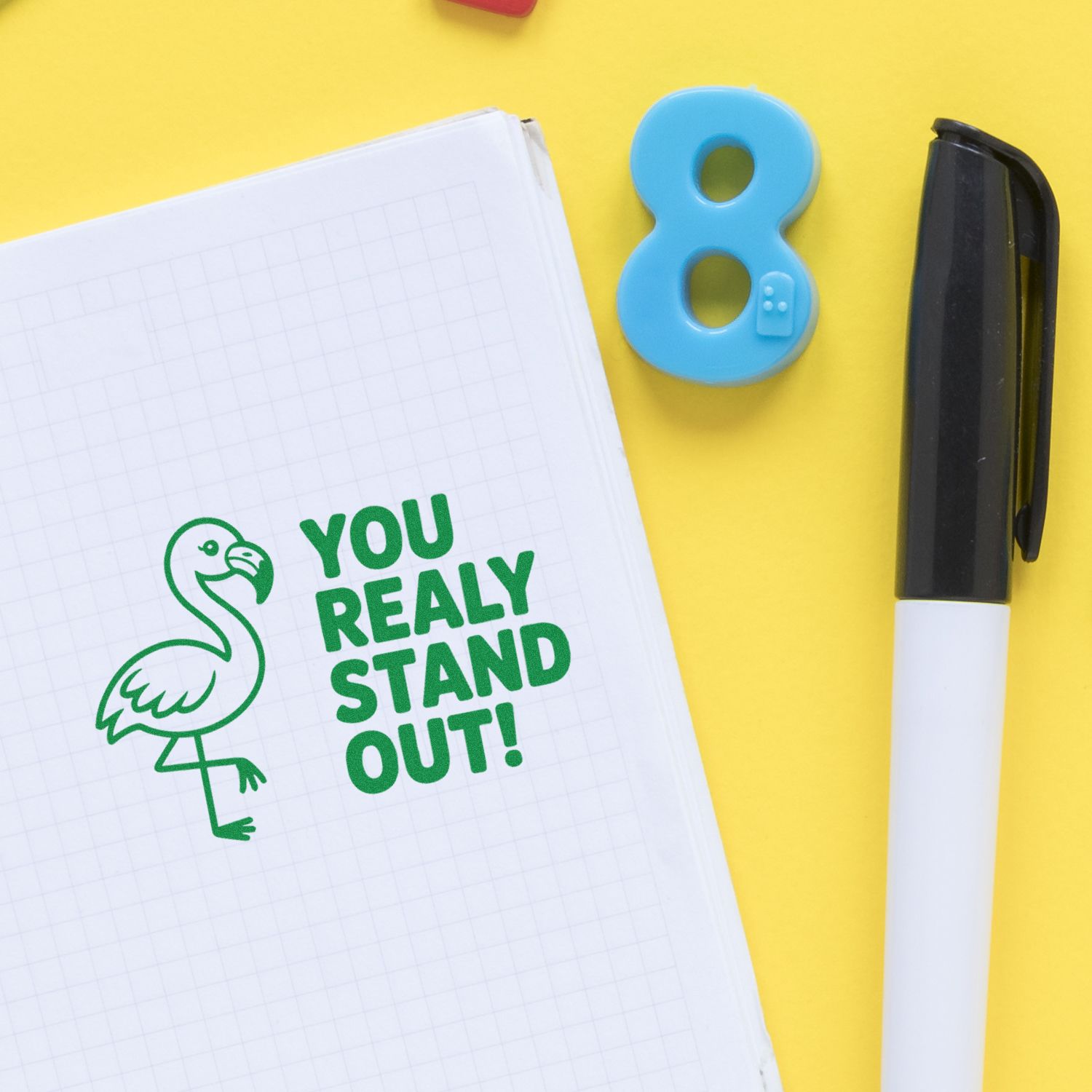 Green flamingo stamp with YOU REALLY STAND OUT! on graph paper, beside a blue number 8 seal and black-and-white pen on yellow.