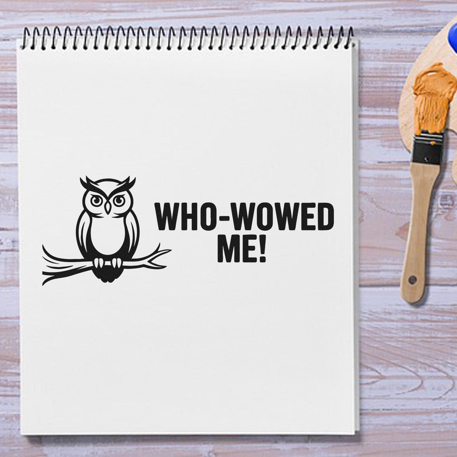 White spiral notebook with black owl stamp and WHO-WOWED ME! seal, beside a paintbrush with orange paint.