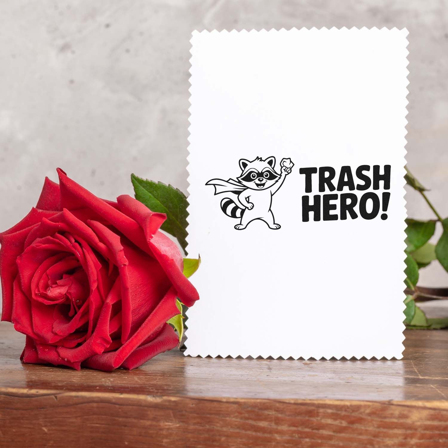 White scalloped stamp and seal card with black raccoon TRASH HERO! design beside a red rose on a wooden table.