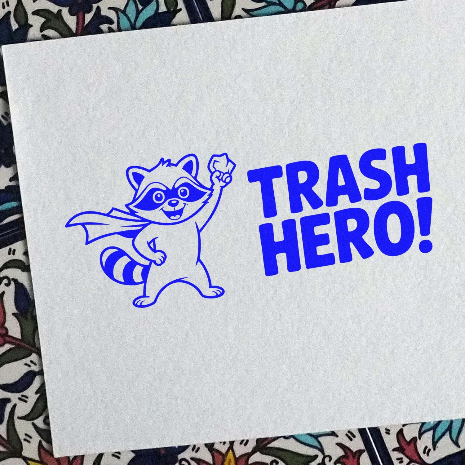 Blue stamp seal of a raccoon superhero with cape and raised fist beside bold TRASH HERO! text on white paper.