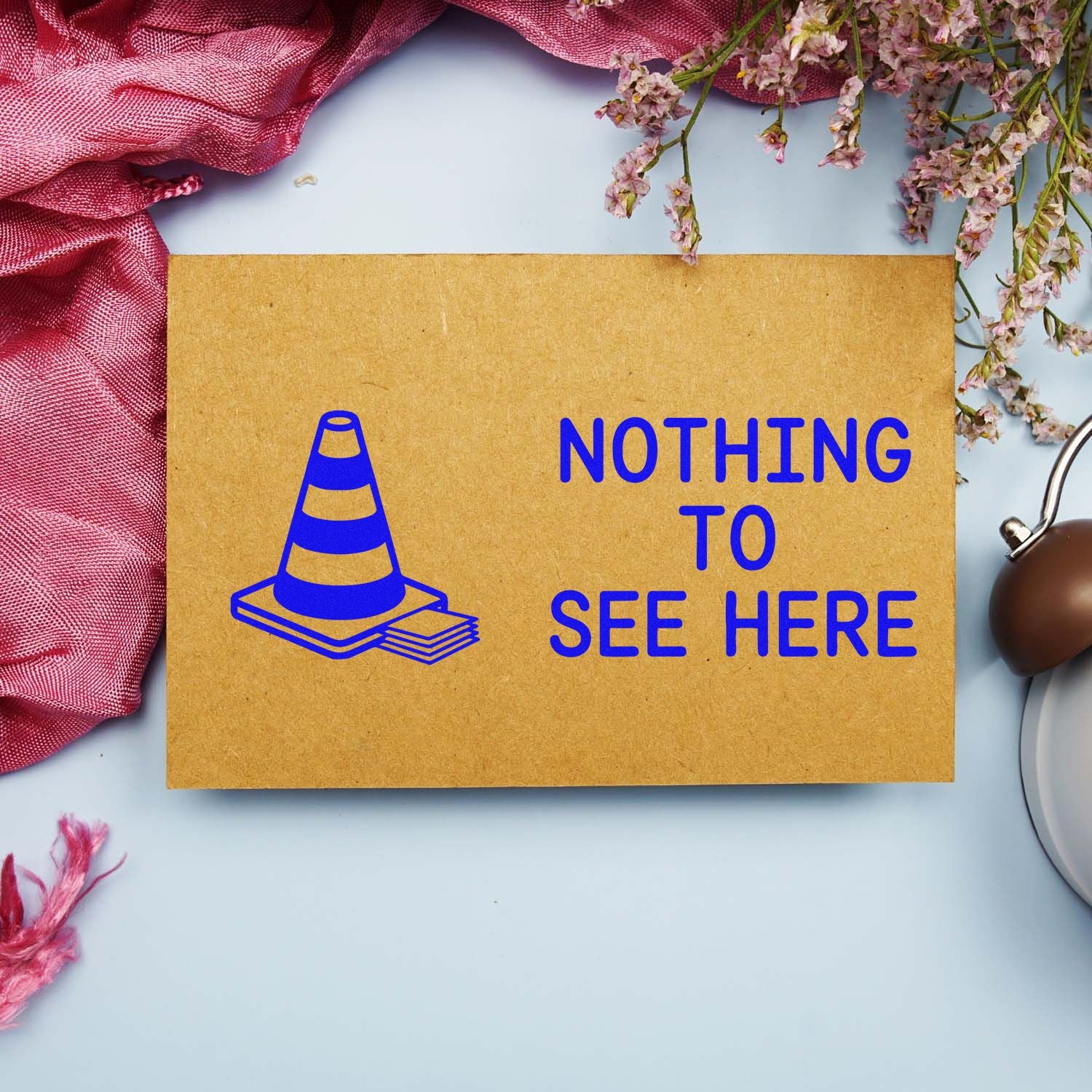 Kraft paper stamp and seal sample with blue cone icon and NOTHING TO SEE HERE, on a light background with pink fabric and flowers.