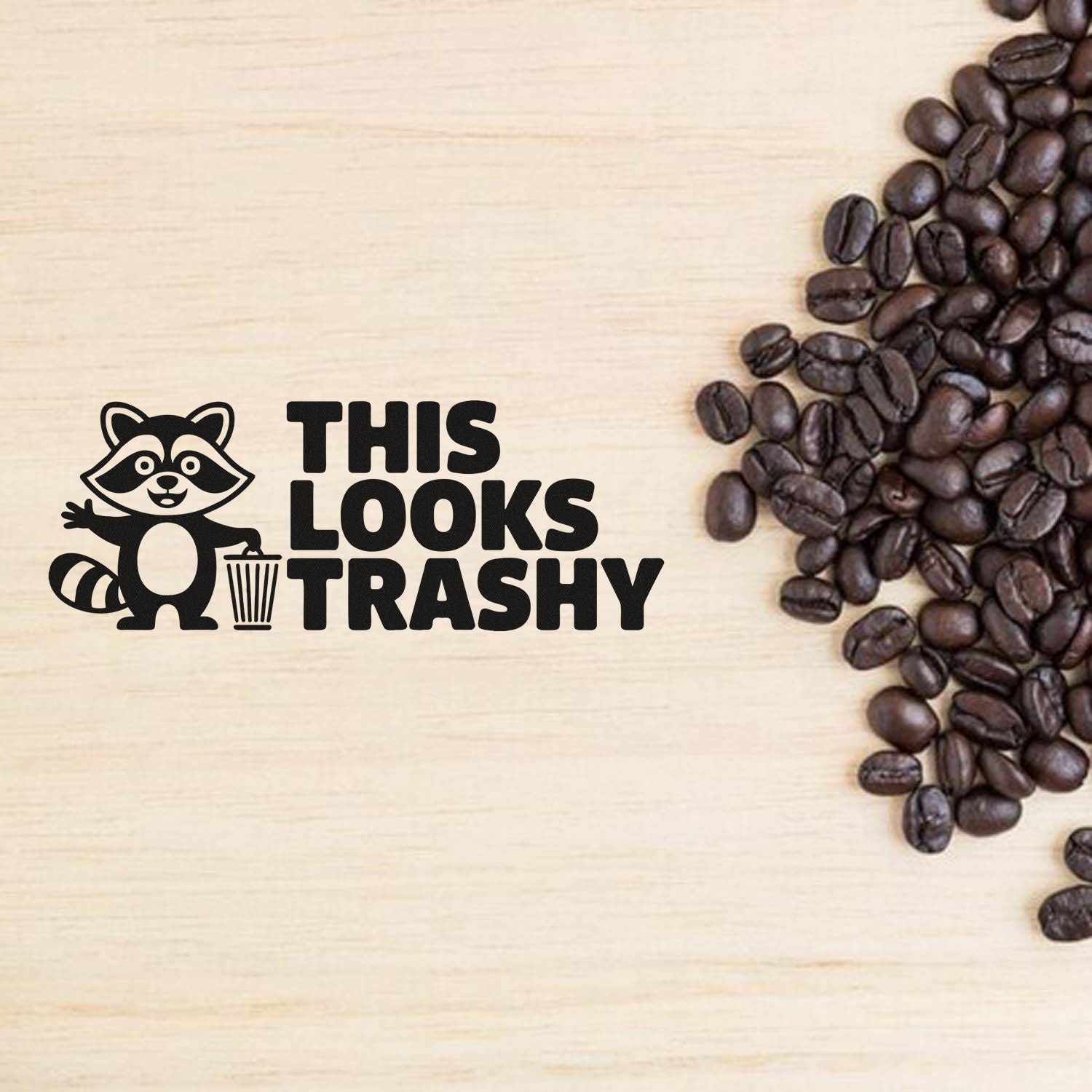 Black raccoon trash stamp and seal reading THIS LOOKS TRASHY, beside coffee beans on a light wood background.