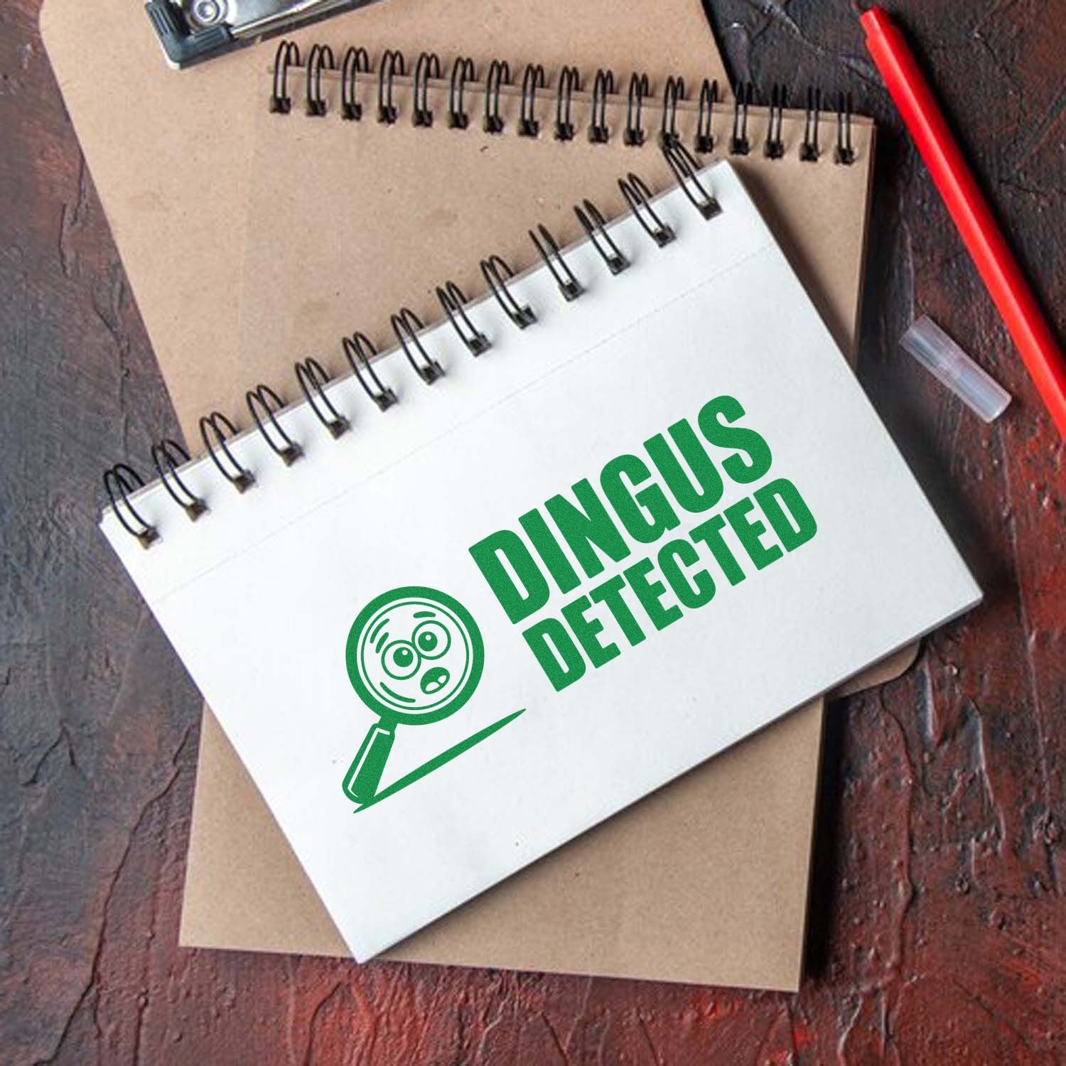 Custom stamp and seal design on a white spiral notebook, green DINGUS DETECTED text with magnifying glass icon, brown pad and red pen.