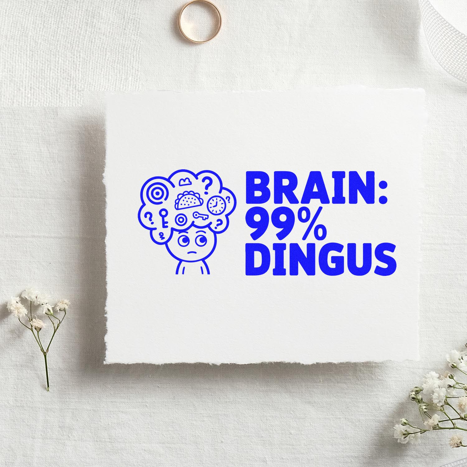 Blue rubber stamp design reading BRAIN: 99% DINGUS with cartoon head on white paper, on a neutral fabric background.