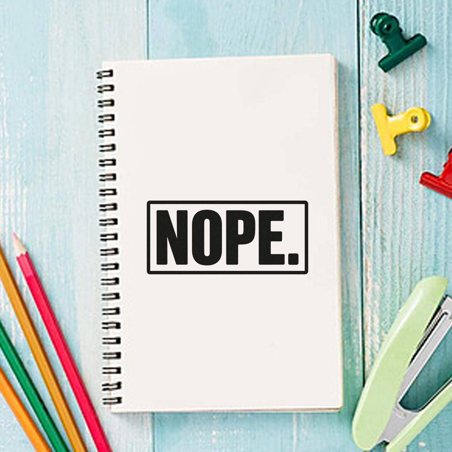Black NOPE stamp on a white spiral notebook, with colorful pencils and binder clips on a light blue wood background.