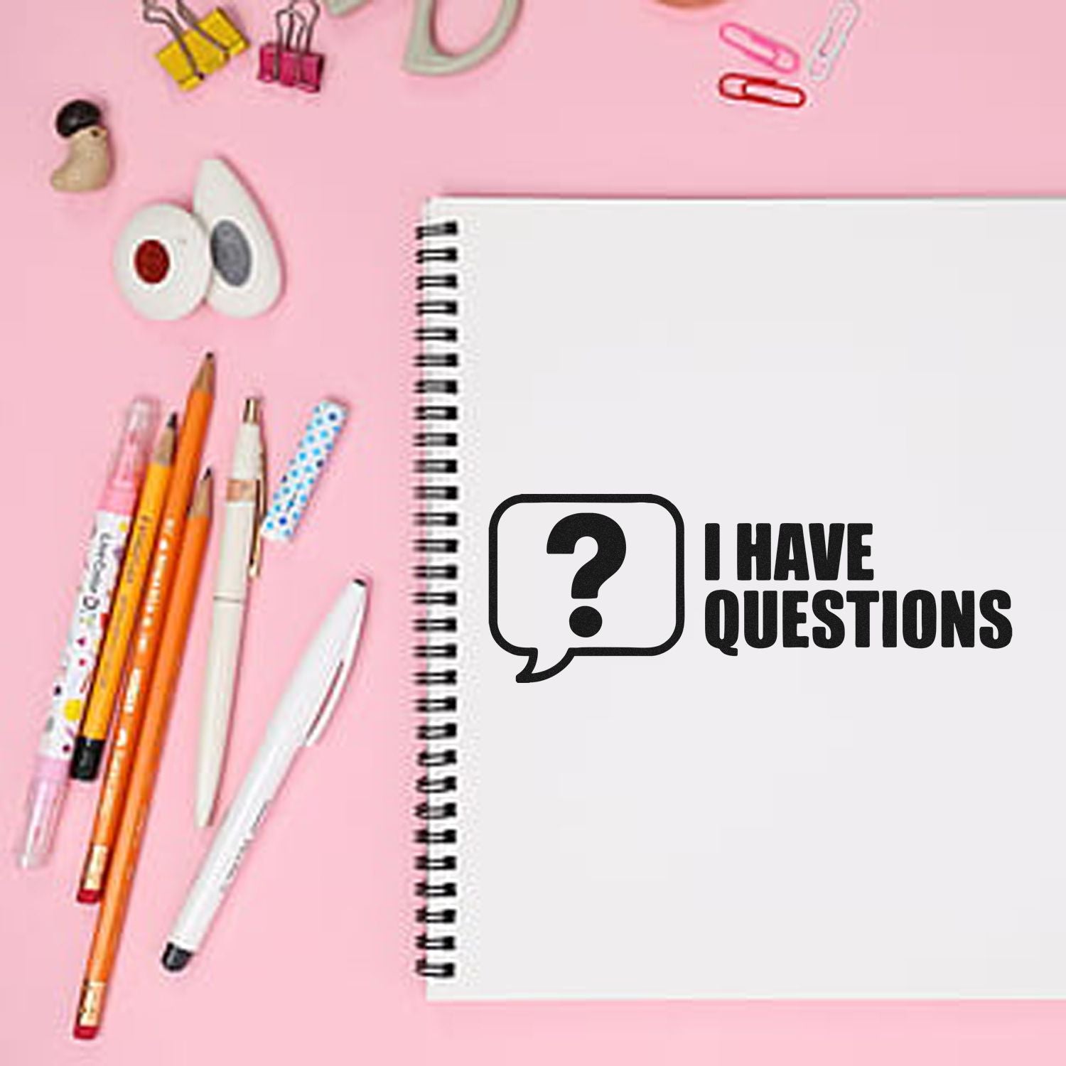 Black question-mark stamp seal on white spiral notebook, pink background, with pens and colorful clips.