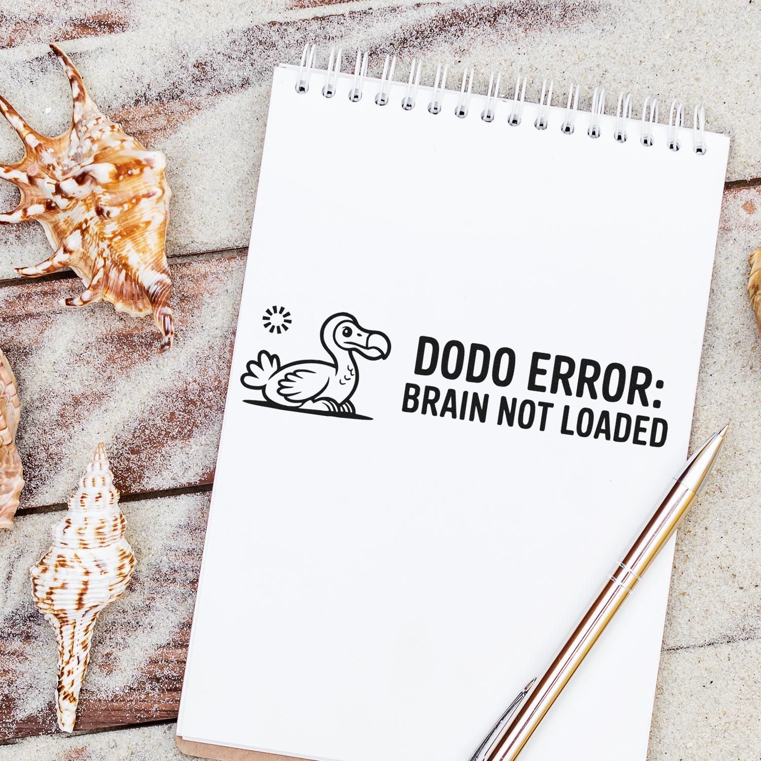 Black dodo stamp on white spiral notebook reads DODO ERROR: BRAIN NOT LOADED, beside seashells and a gold pen.