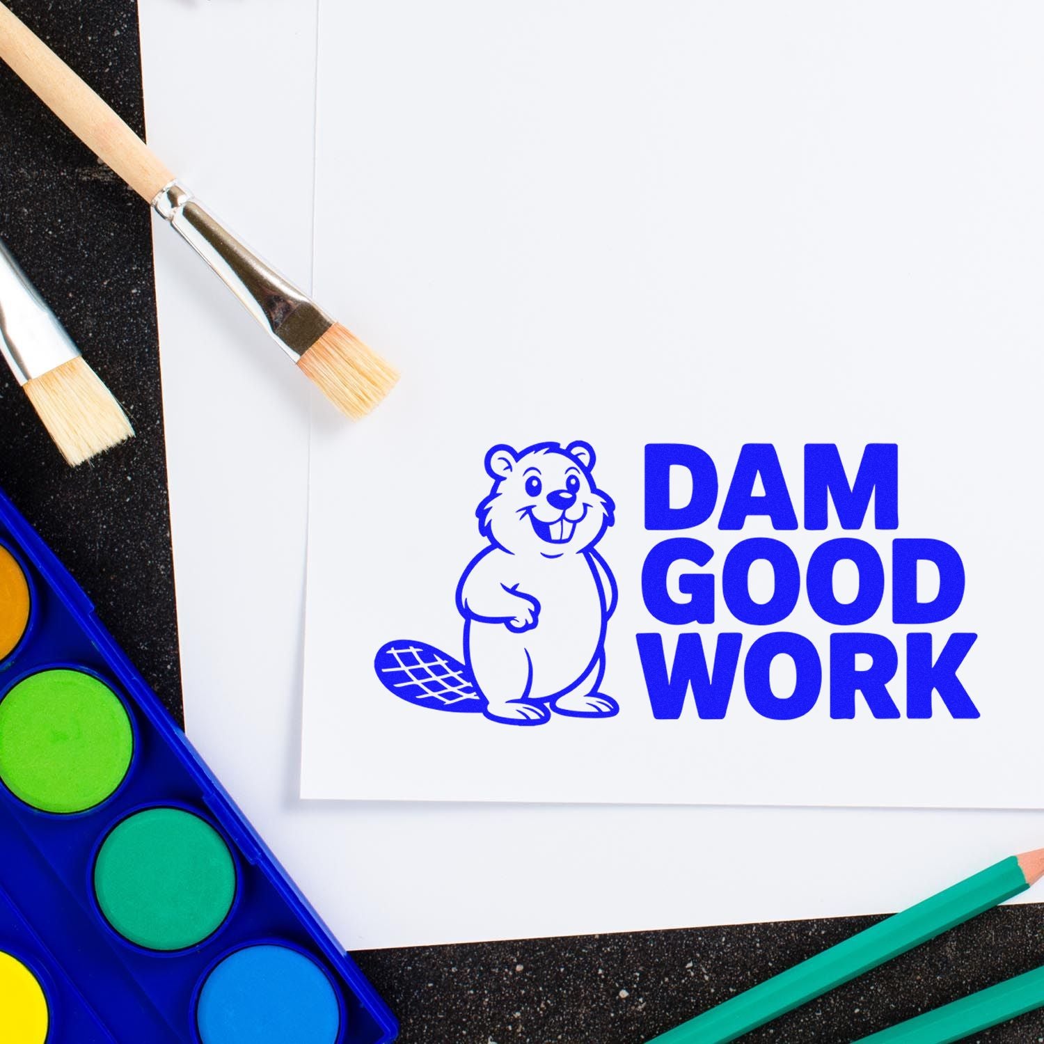 Blue beaver DAM GOOD WORK stamp and seal design on white paper, with paintbrushes, watercolor palette, and colored pencils.