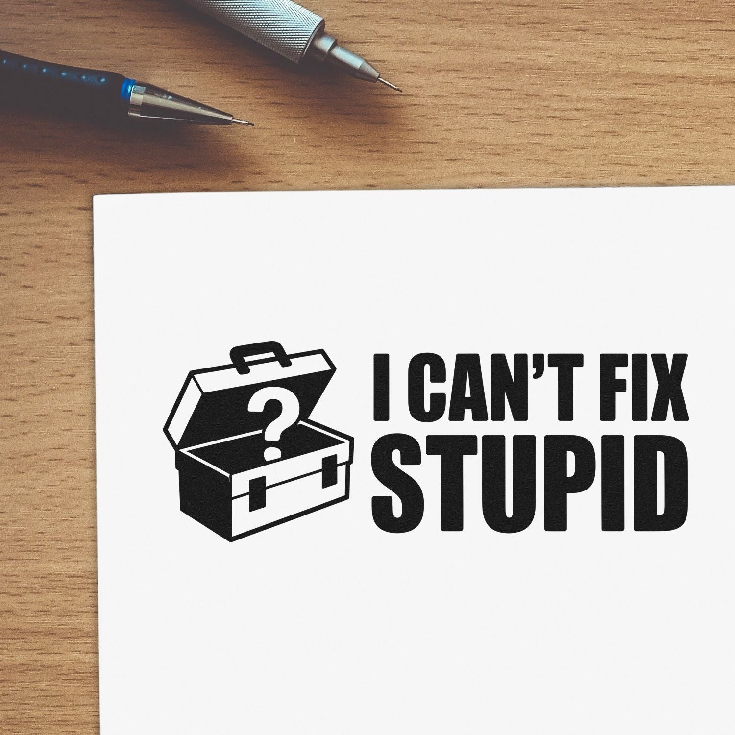 Black stamp and seal design with toolbox question mark and bold I CANT FIX STUPID text on white paper beside pens.
