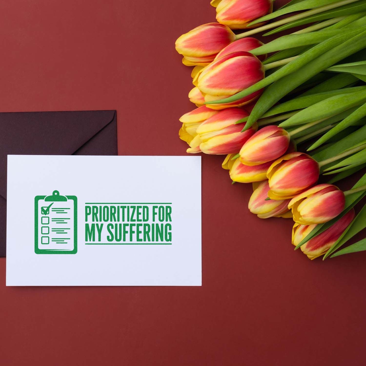 Green rubber stamp seal reading Prioritized for My Suffering on white card beside pink-yellow tulips and dark envelope.