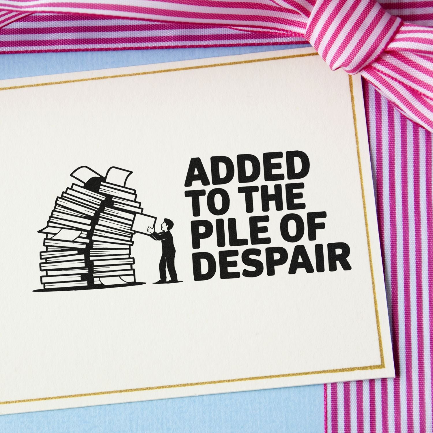 Black stamp seal reading Added to the Pile of Despair with paper stack icon on white card, gold border and pink striped ribbon.