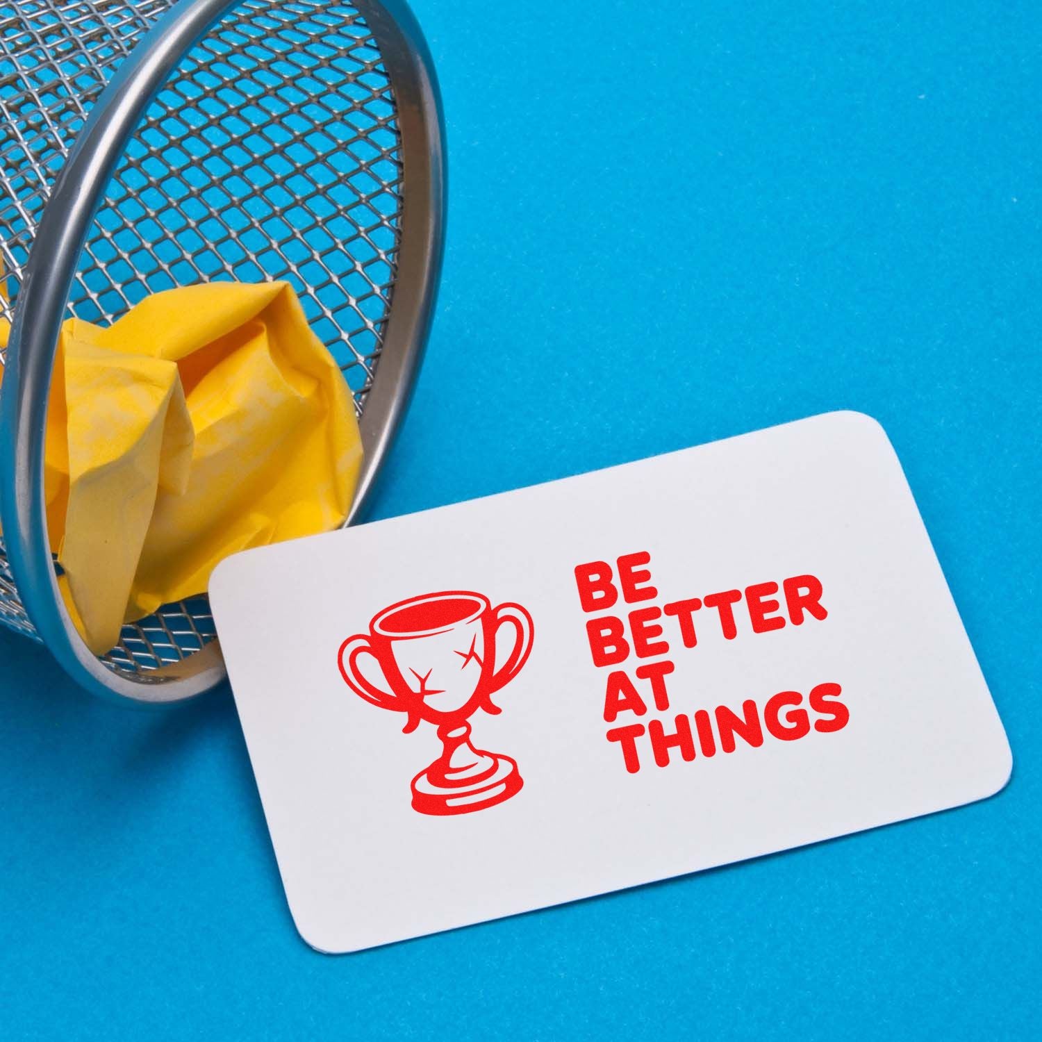 Red trophy stamp/seal on white card with Be Better at Things, beside a metal mesh basket and yellow cloth on blue background