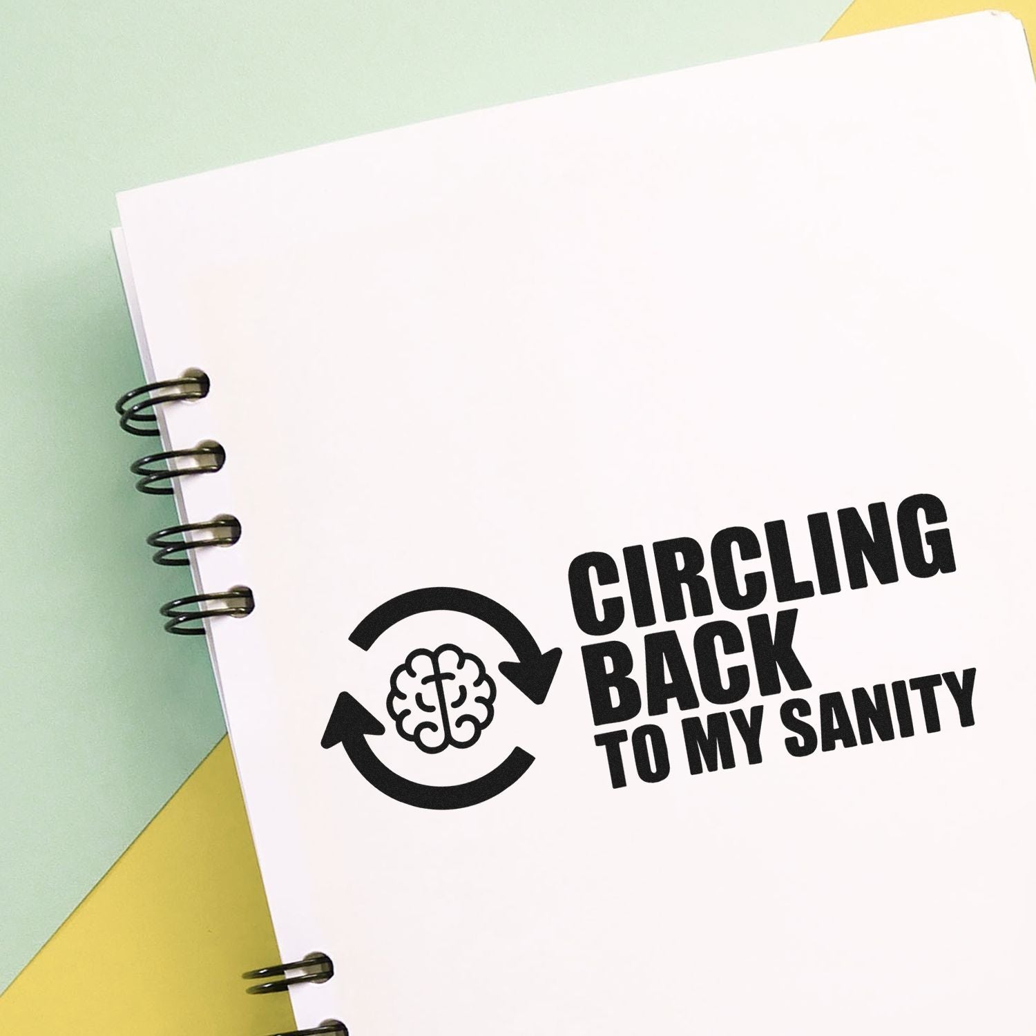 Black stamp-style seal with brain in circular arrows and Circling back to my sanity on white notebook, pastel green and yellow.