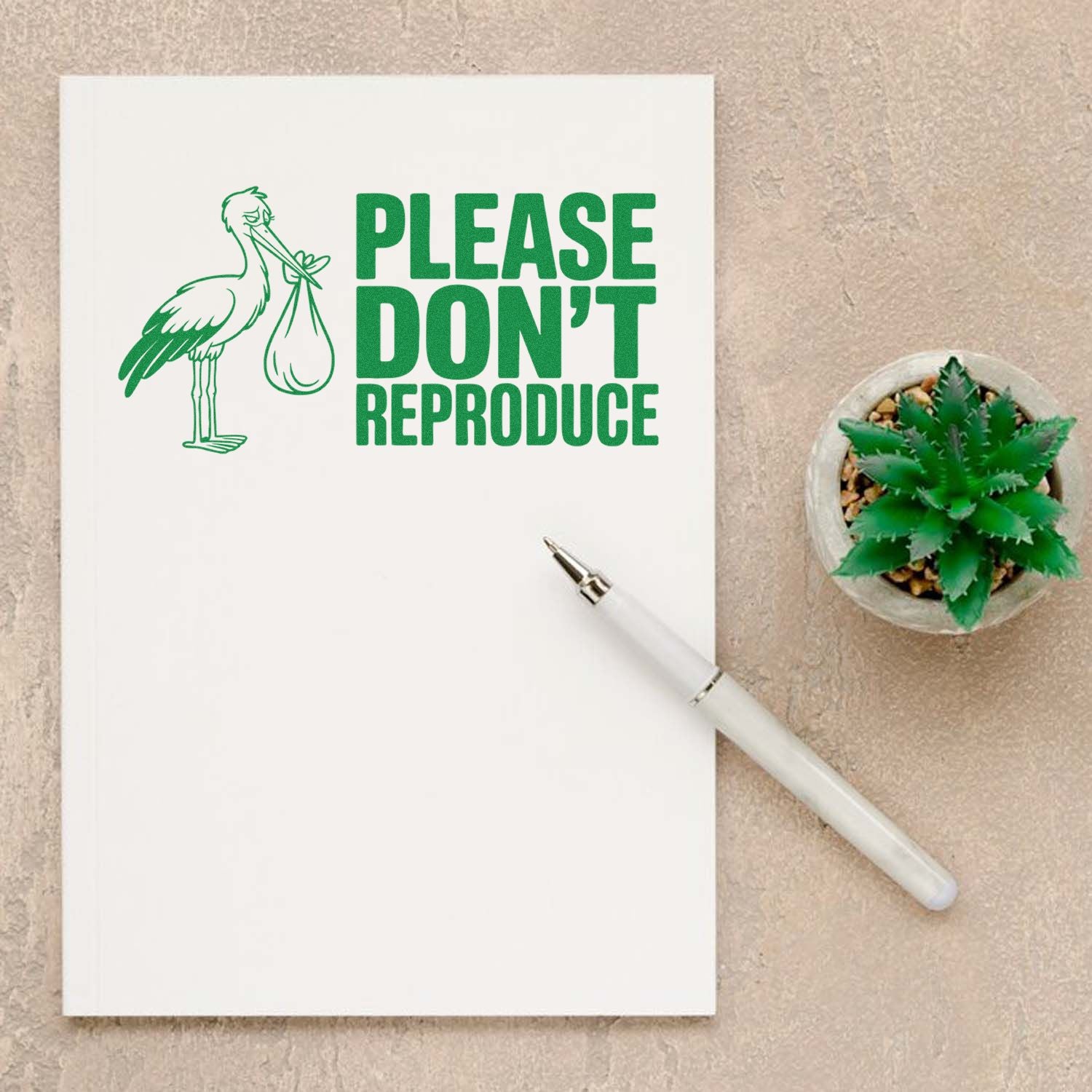 Green Please Dont Reproduce stamp seal with stork carrying bundle on white notebook, beside pen and succulent.