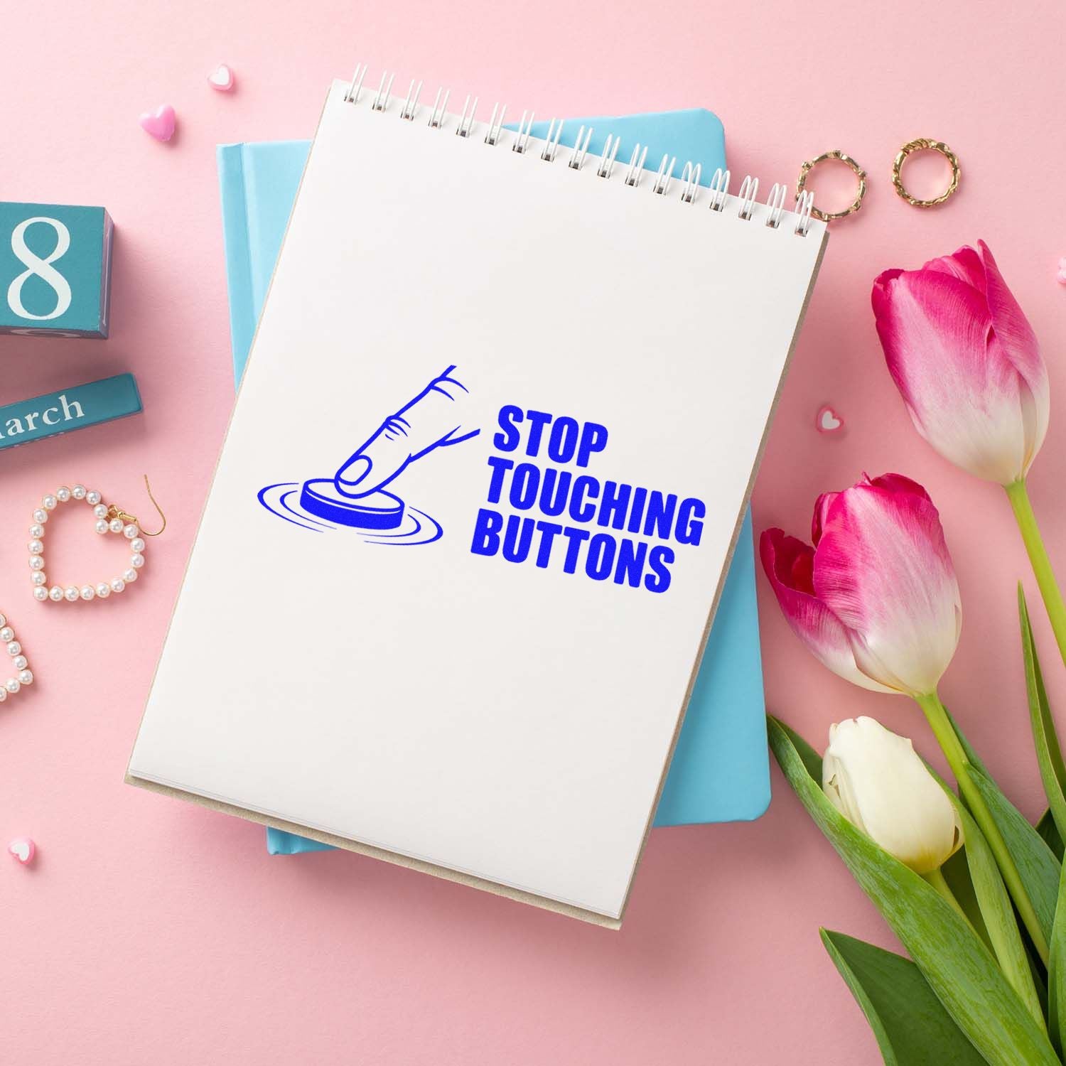 Blue self-inking stamp/seal reading STOP TOUCHING BUTTONS on a white notebook, with pink tulips and pastel accessories.