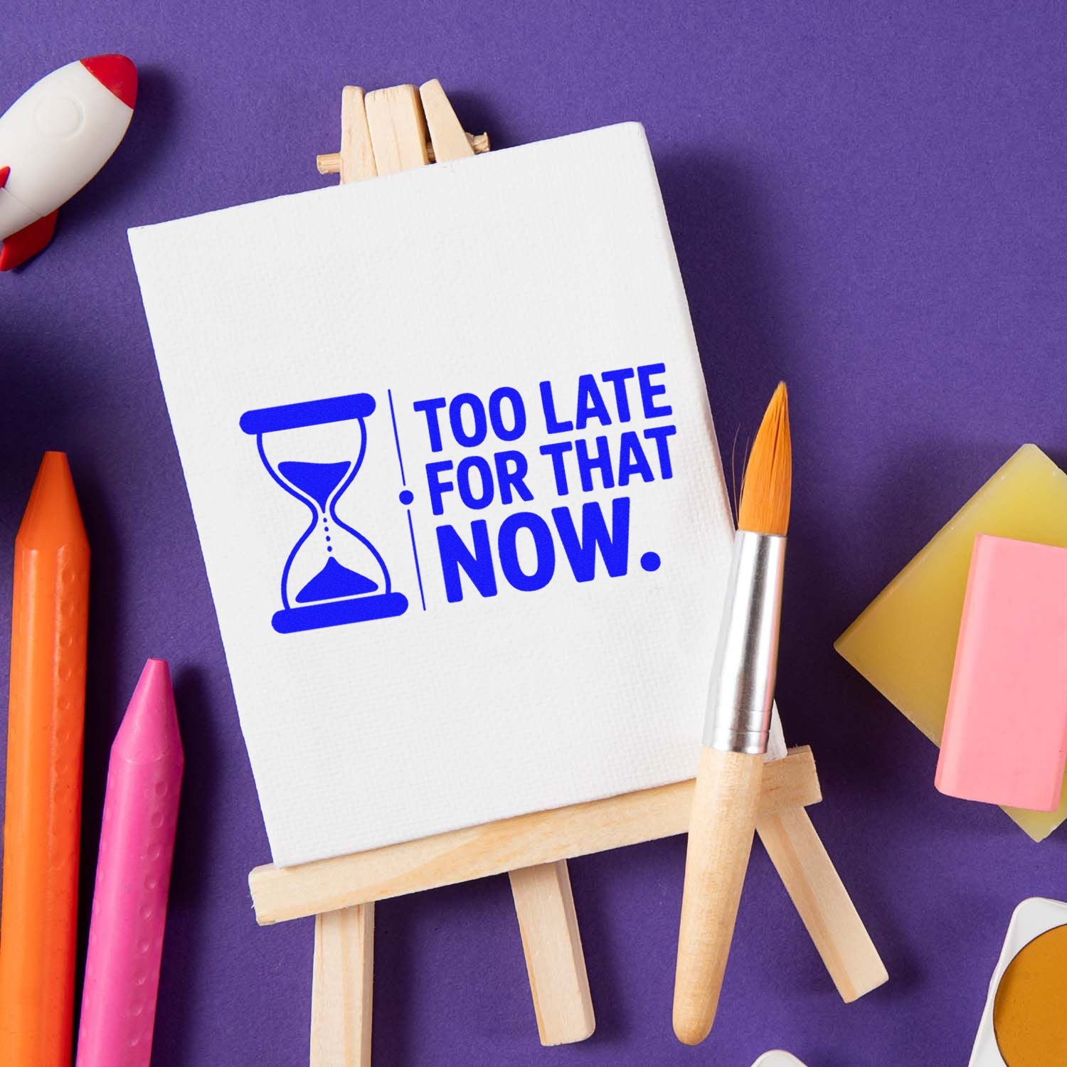 Blue hourglass rubber stamp reading Too Late for That Now on white easel, with colorful art supplies on a purple background.