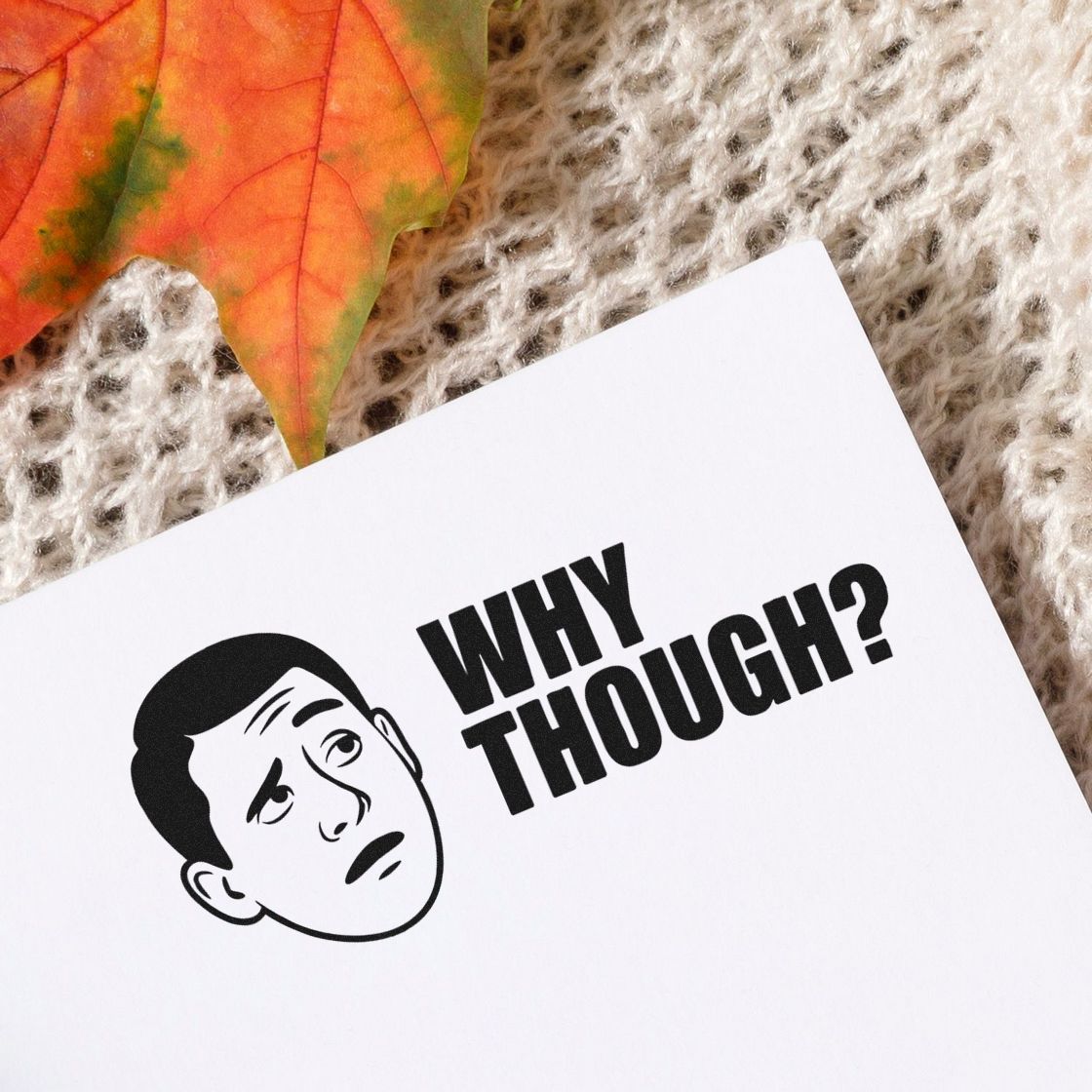 Black Why Though? stamp and seal with puzzled face on white paper, beside orange autumn leaf on beige knit fabric.
