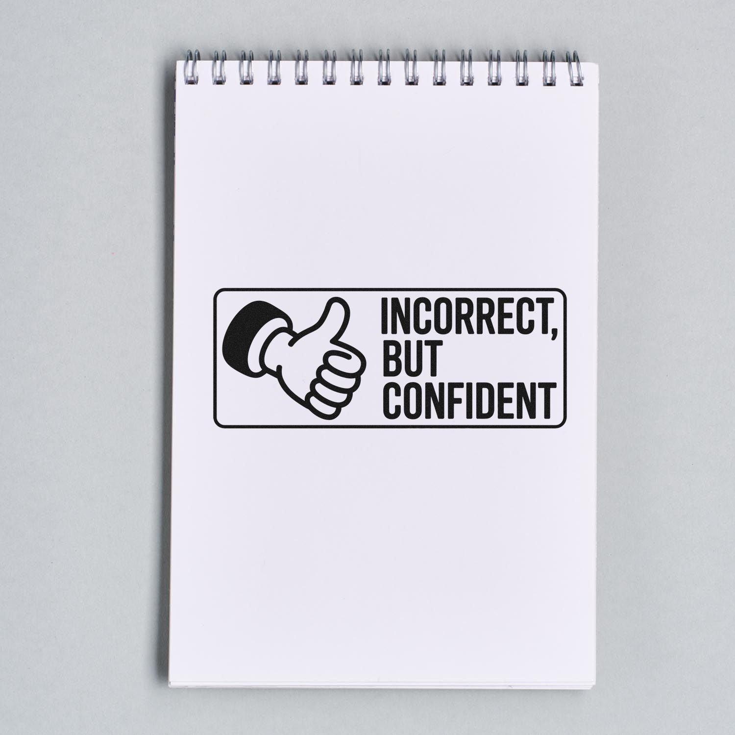 Black self-inking stamp seal with thumbs-up icon and Incorrect, but confident on a white spiral notepad.