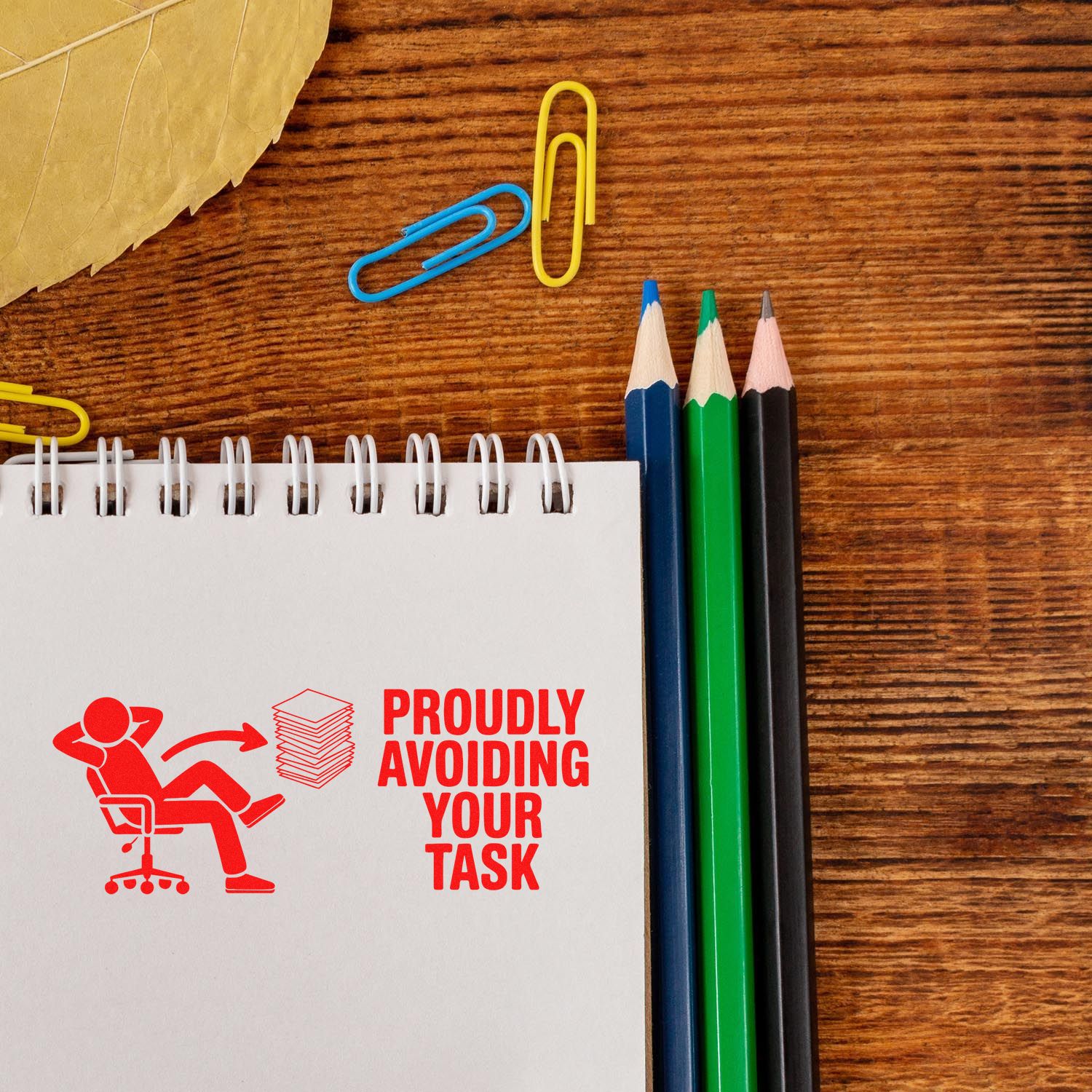 Red self-inking stamp and seal design on notebook reads Proudly avoiding your task, with pencils and paperclips on wood.