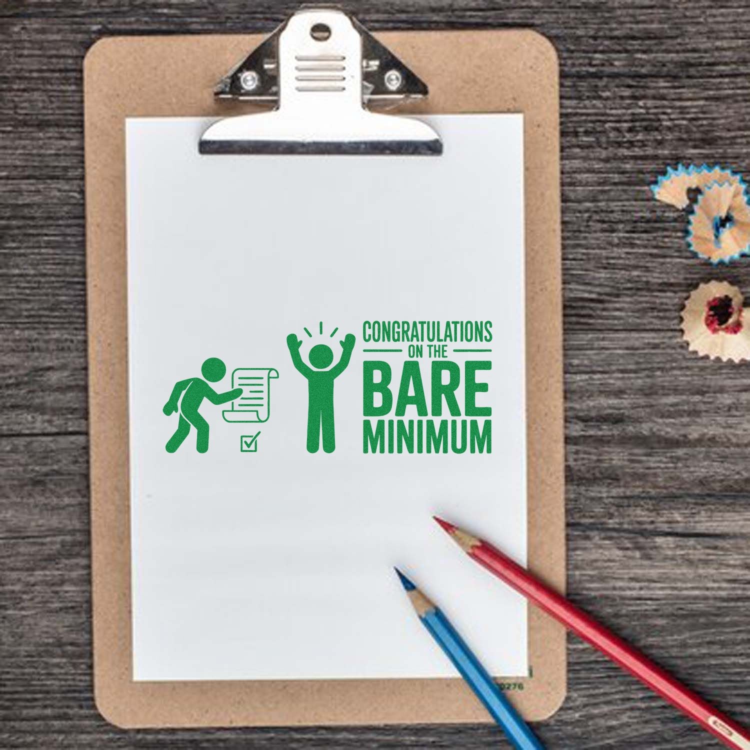 Green rubber stamp and seal design on clipboard paper reading Congratulations on the Bare Minimum, with red and blue pencils on a wood desk.