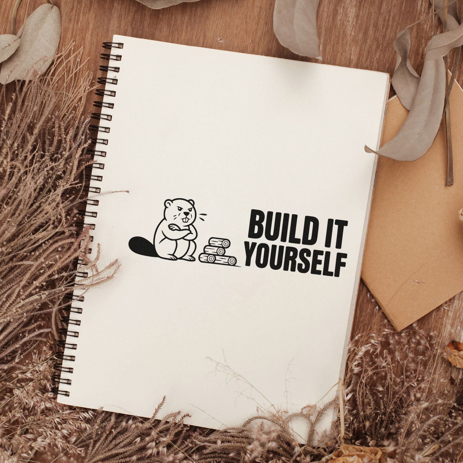 Black stamp seal with beaver and Build It Yourself on a cream spiral notebook, surrounded by dried beige foliage.