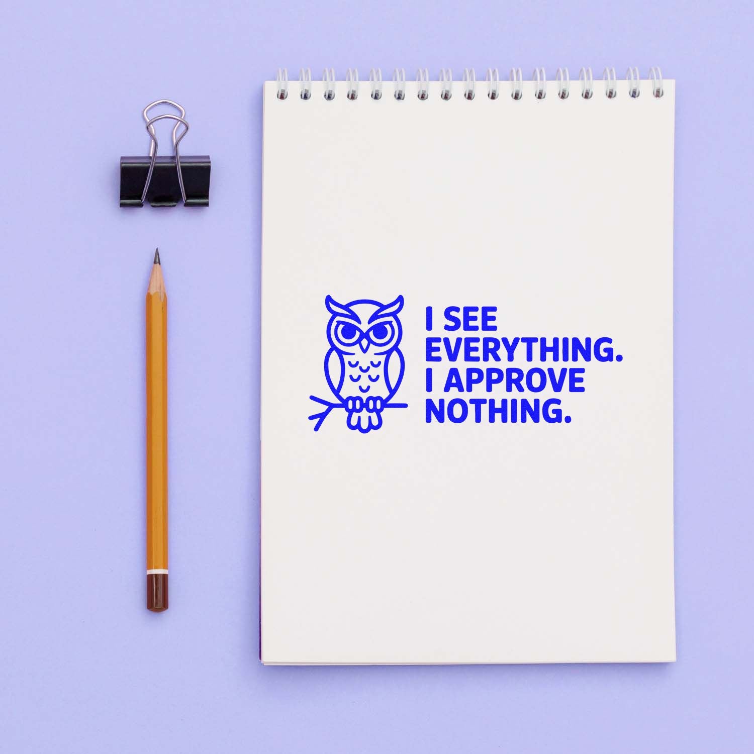 White spiral notebook with a blue owl rubber stamp reading "I SEE EVERYTHING. I APPROVE NOTHING," beside a pencil and binder clip.