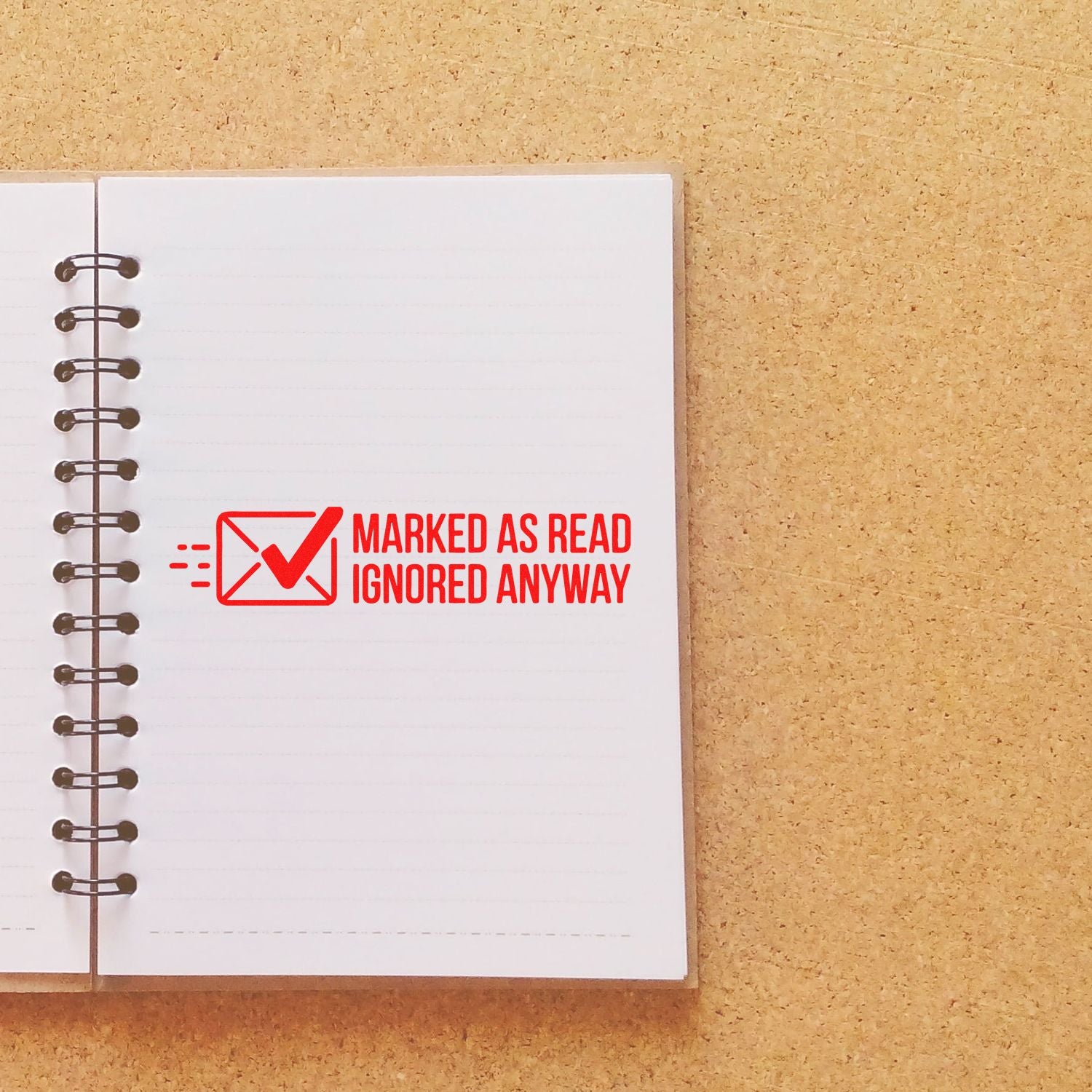 Red rubber stamp seal reading Marked as read ignored anyway on a white spiral notebook over a tan cork background.